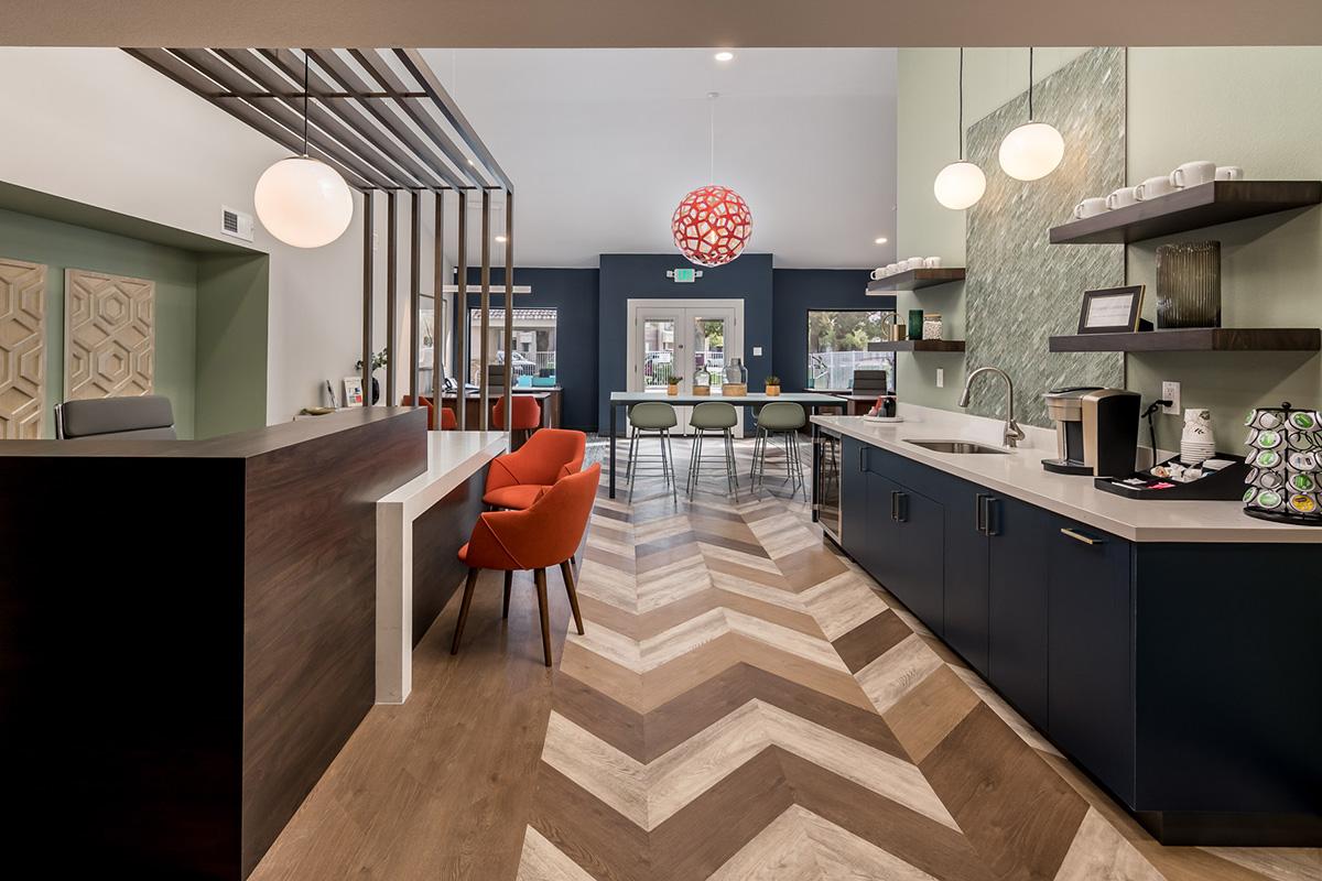 Modern interior of a reception area featuring a stylish wooden desk, bright orange chairs, and a bar-style seating arrangement. The chevron-patterned floor complements the green and blue walls, with decorative elements like a large pendant light and a coffee station visible in the background.