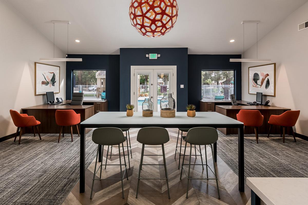 A modern office space featuring a central table with four chairs, surrounded by two workstations with orange chairs. Large windows provide natural light, and decorative art is displayed on the walls. A unique pendant light hangs from the ceiling, creating a contemporary atmosphere.
