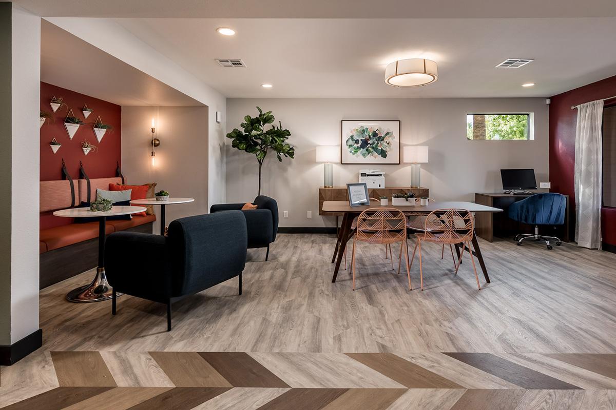 A modern, inviting interior space featuring a seating area with a round table and chairs, a potted plant, and stylish art on the walls. The wood flooring contrasts with the colorful decor, creating a warm atmosphere. A desk area is visible in the back, equipped with a computer and chair, enhancing the functional design.