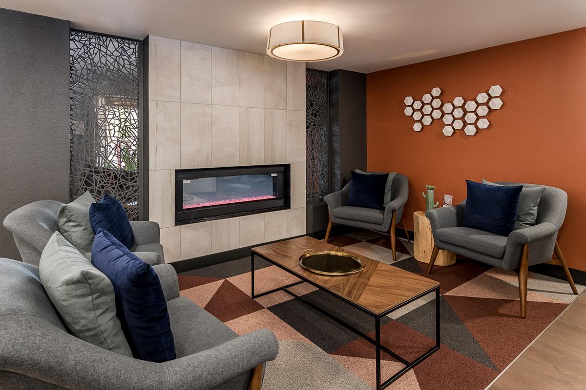 A modern living room featuring two comfortable gray chairs with blue cushions, a stylish coffee table with a decorative bowl, a cozy fireplace surrounded by neutral tiles, and an accent wall in orange adorned with hexagonal wall art. The floor is covered in a geometric rug.