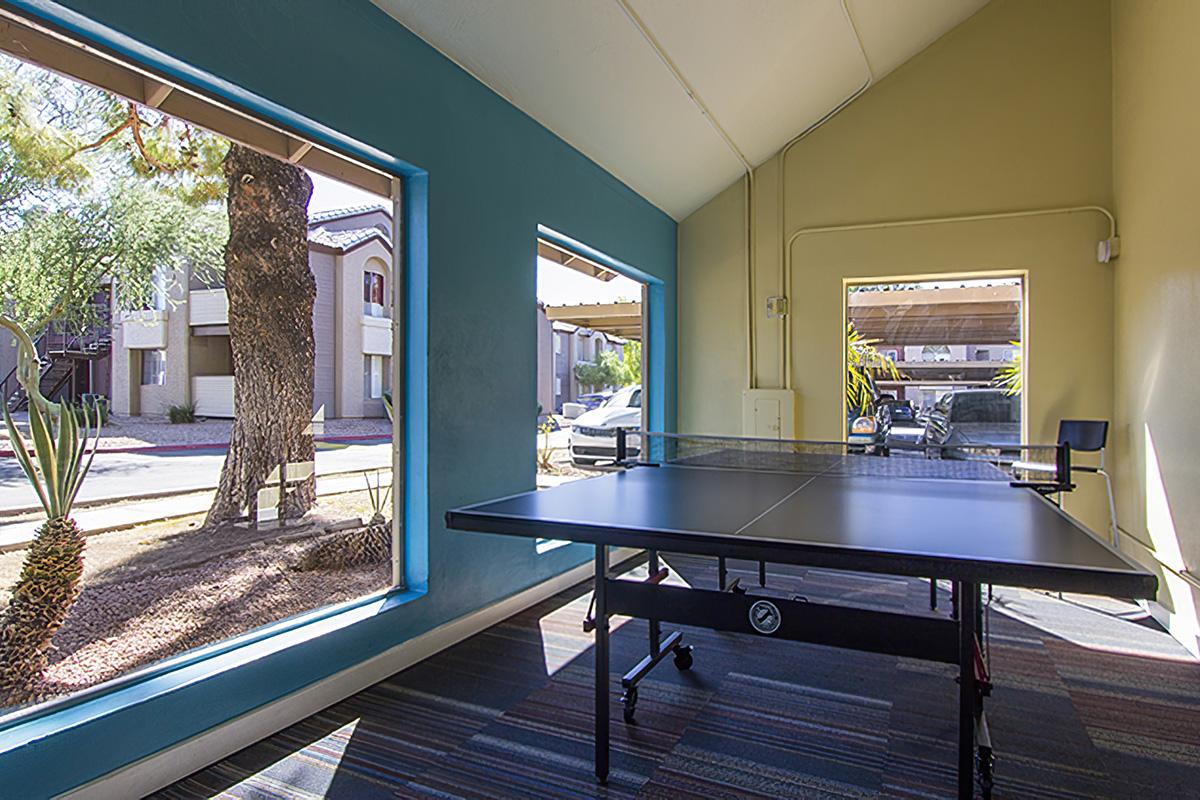 A bright indoor area with large windows showcasing a view of the outdoors. A black ping pong table is centered in the space, surrounded by colorful walls. The area appears well-lit and inviting, with a glimpse of greenery and parked cars outside.