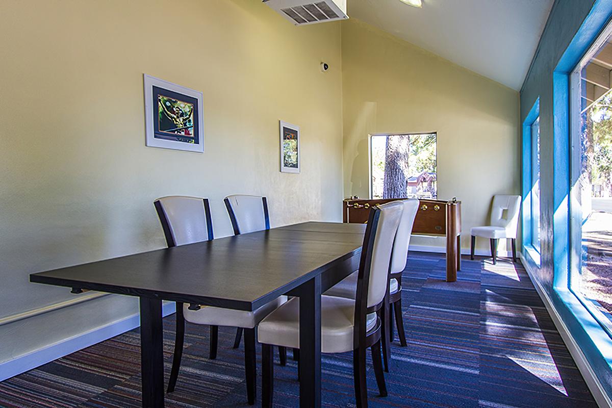 A well-lit meeting room featuring a long black table surrounded by light-colored chairs. The walls are painted a soft yellow, and there are framed artworks on the walls. A large window allows natural light to brighten the space, and there is a wooden cabinet visible in the background. The floor has a striped carpet.
