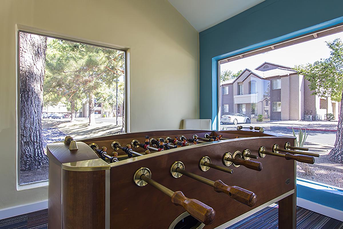 A foosball table is positioned in a sunny room with large windows. Outside, trees and a residential building can be seen. The room features light-colored walls and a teal accent wall, creating a vibrant atmosphere for leisure activities.
