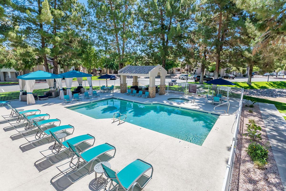 A bright outdoor pool area surrounded by green trees. There are several lounge chairs arranged around the pool, and two shaded cabanas for seating. An outdoor gazebo and umbrellas provide additional shade, creating a relaxing atmosphere for guests.