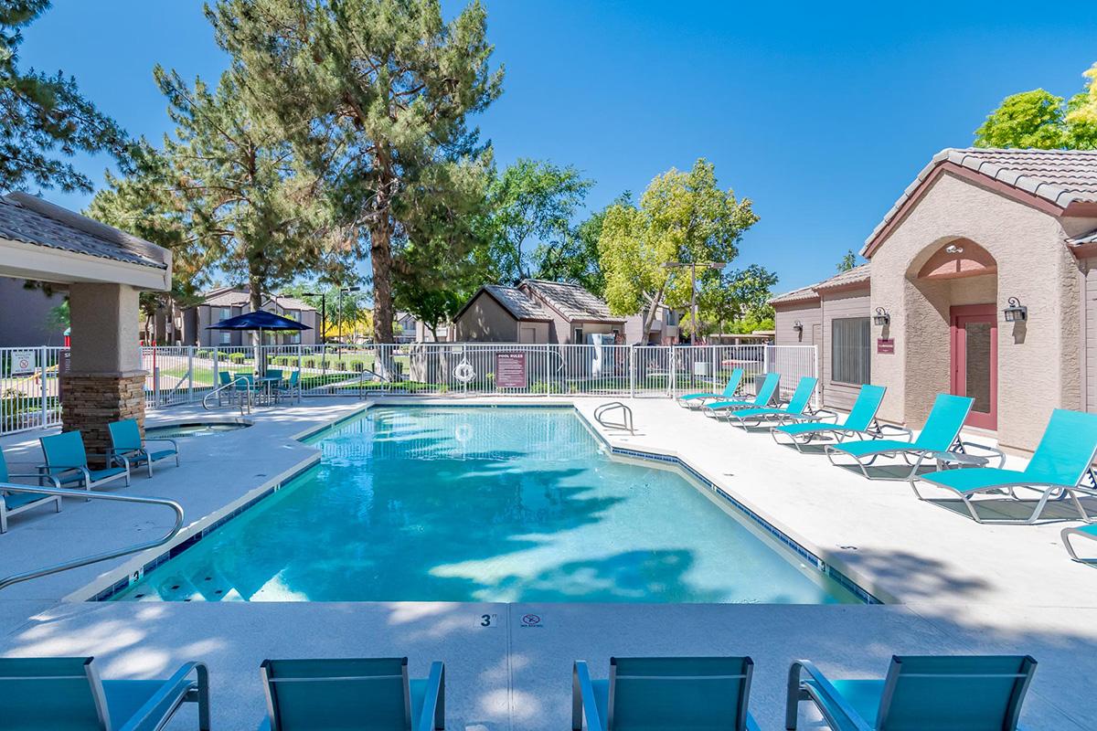 A bright outdoor pool area surrounded by lounge chairs and trees. The pool is clear and inviting, with a white deck and a fence in the background. Several buildings with a suburban style are visible nearby, set against a clear blue sky.