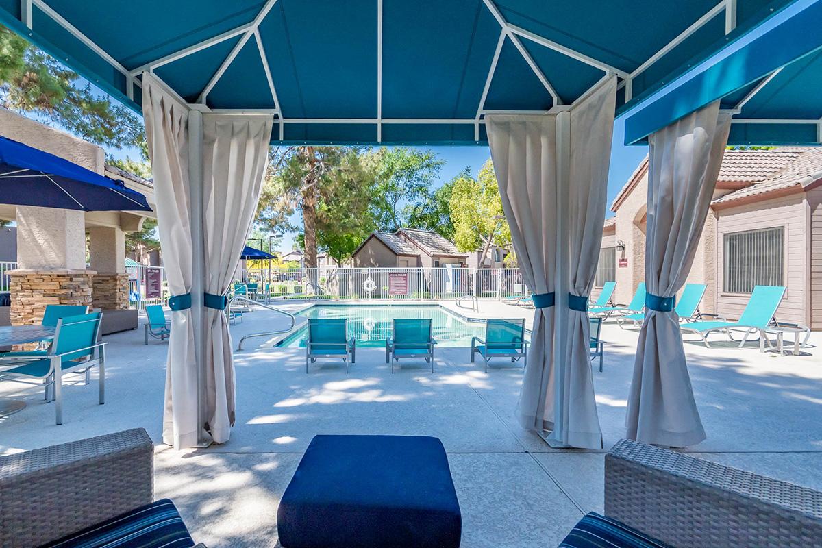 View from a shaded cabana overlooking a swimming pool area with lounge chairs, surrounded by trees and buildings. The scene features white drapes gently blowing in the breeze, creating a relaxing outdoor space perfect for leisure. Sunlight filters through, enhancing the inviting atmosphere.