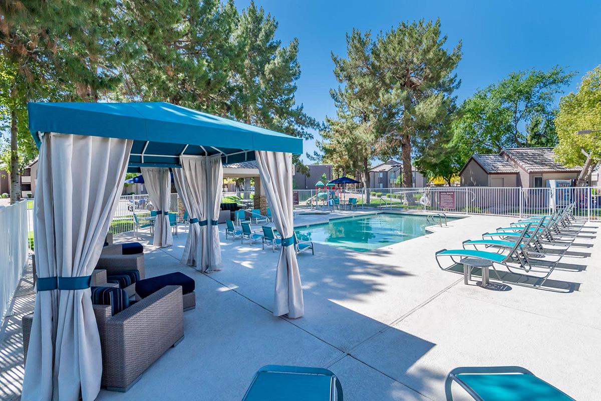 A bright and inviting pool area featuring a swimming pool surrounded by lounge chairs. A shaded cabana with curtains provides a relaxing seating space, while green trees and colorful decor enhance the outdoor atmosphere. The setting is perfect for leisure and enjoyment.