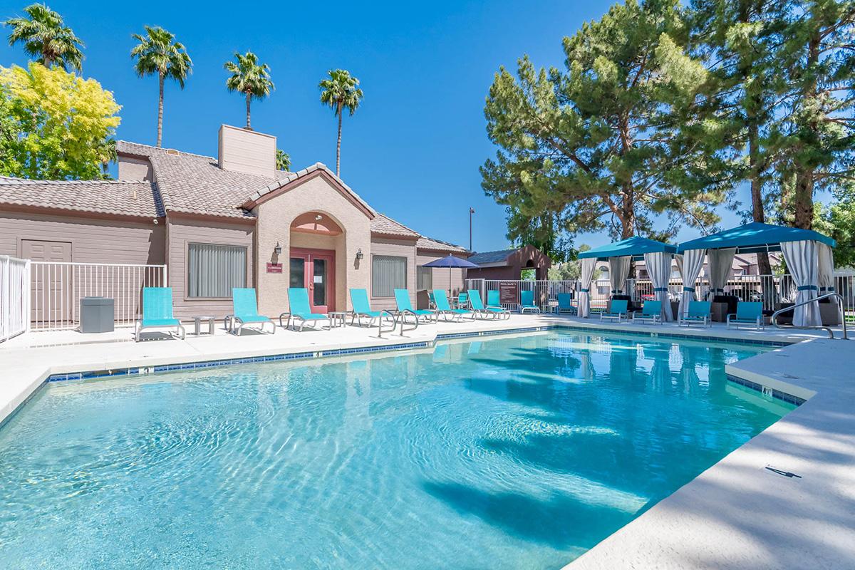 A sunny outdoor pool area featuring a clear blue water pool surrounded by lounge chairs and shaded cabanas. Palm trees provide greenery in the background, and a spacious building with large windows is visible nearby. The atmosphere is inviting and relaxing, perfect for leisure.