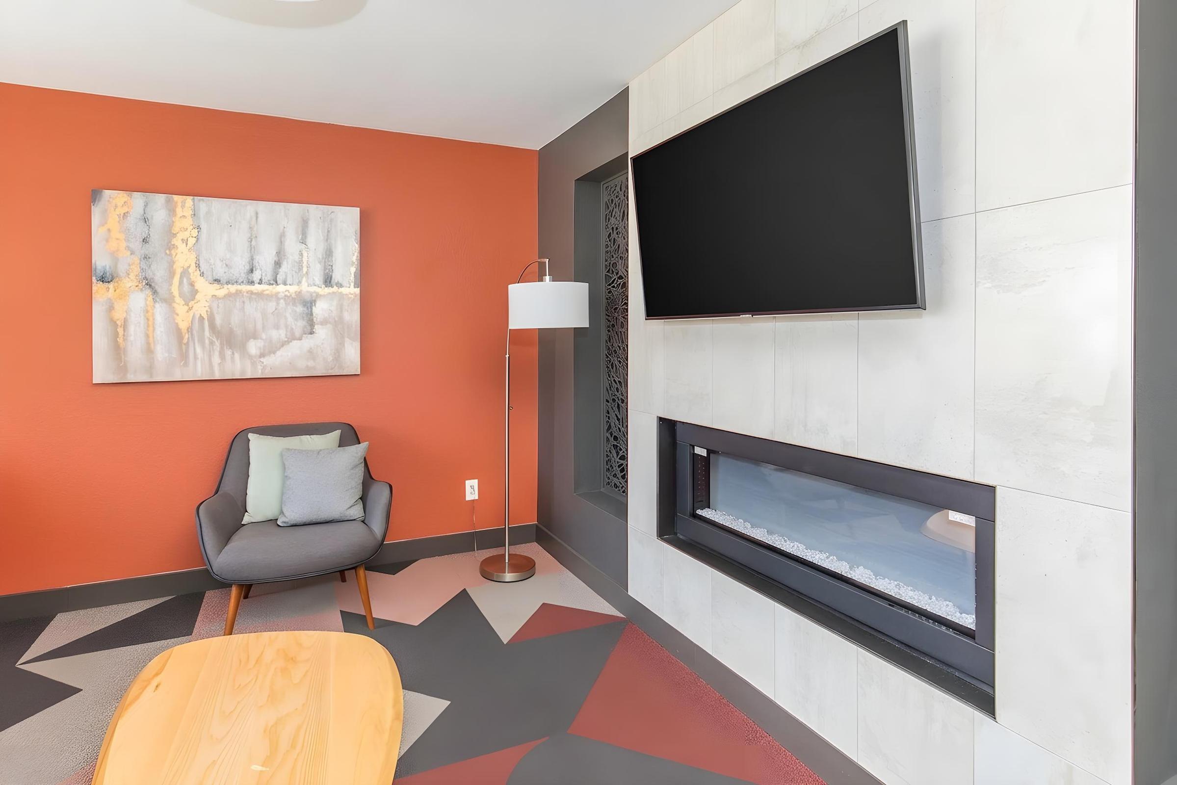 A modern living room features an orange accent wall, a stylish gray armchair with decorative pillows, a sleek floor lamp, and a mounted flat-screen TV. The wall includes a contemporary fireplace with a minimalist design, and the floor is adorned with geometric patterns in muted colors.