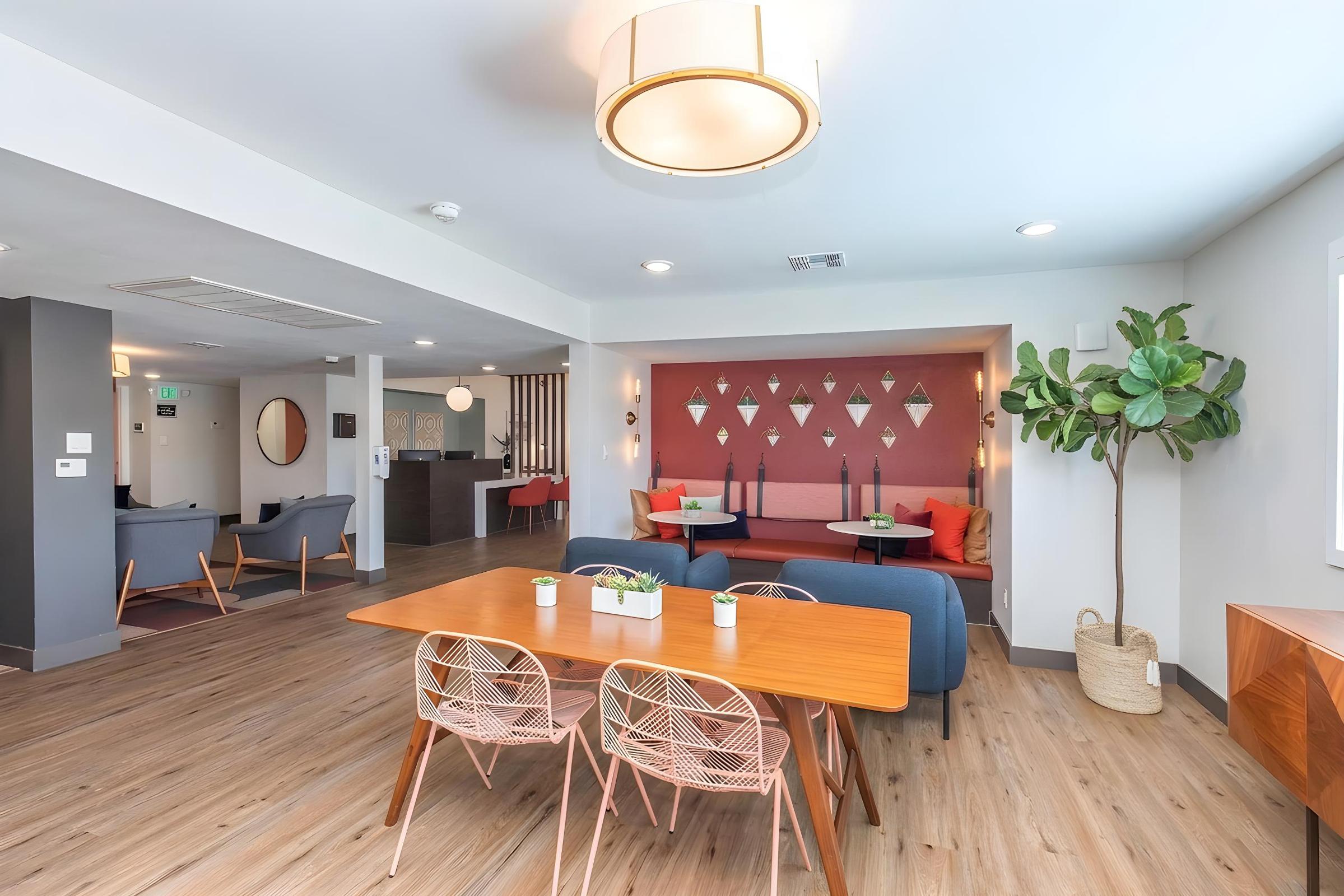 Modern interior of a cozy lounge area featuring a wooden dining table with chairs, a blue sofa, and a leafy plant. The walls are decorated with geometric art, and there are bright accents in red and orange. Ample natural light enhances the welcoming atmosphere, making it a pleasant social space.