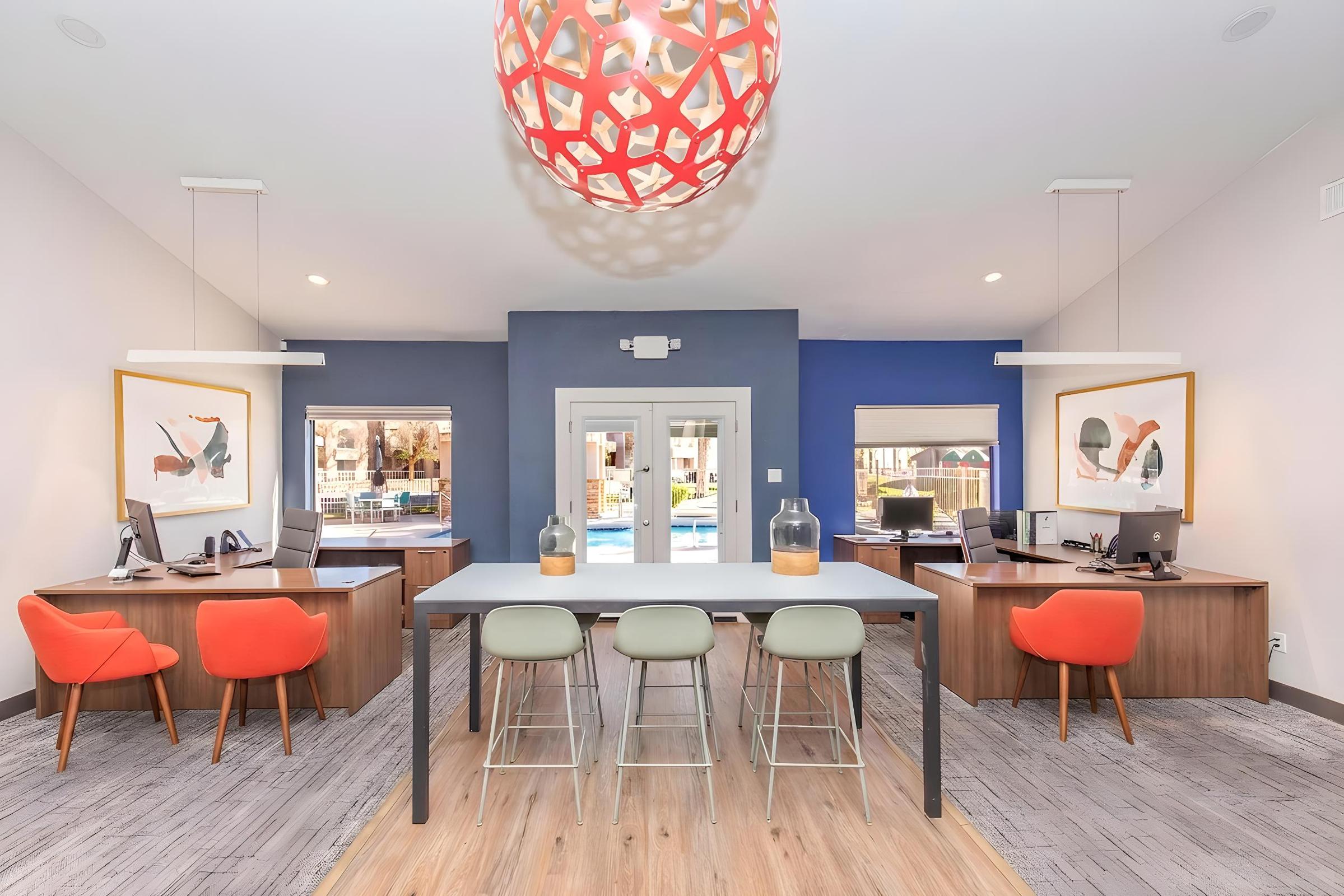 Modern office reception area with two desks featuring computers and office chairs, surrounded by colorful wall art. A large, round pendant light hangs from the ceiling, and there are stylish barstools at a central table. Large windows allow natural light, and a swimming pool is visible outside.