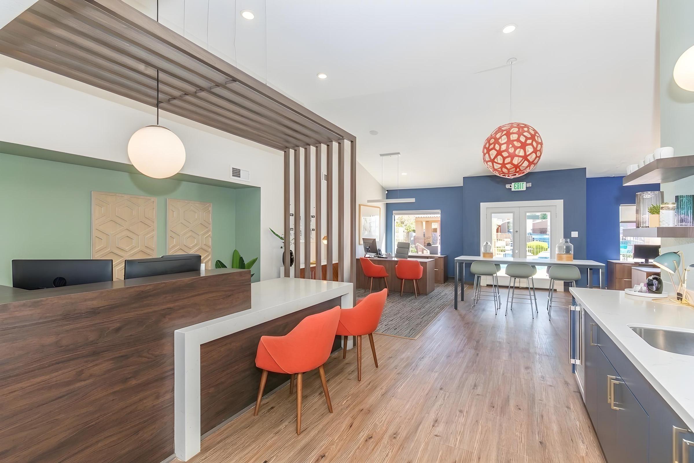 Interior of a modern office reception area featuring a welcoming front desk, colorful orange chairs, and bright lighting. The space has green and wood accents, a large window allowing natural light, and a casual seating area for visitors.
