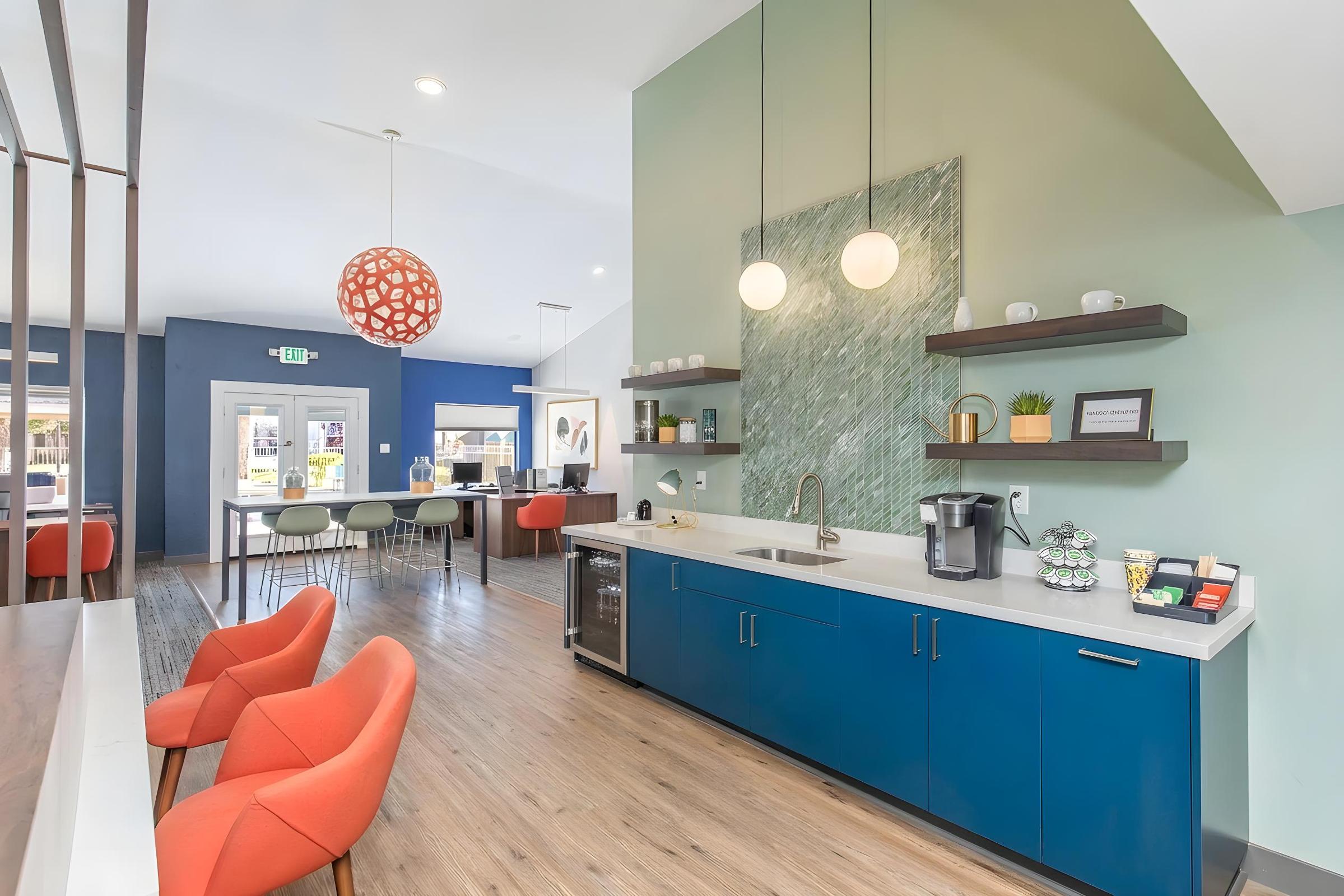 Modern interior of a communal kitchen space featuring a blue kitchen cabinet with a sink, coffee maker, and snacks. Cozy orange chairs are arranged around a central table, while decorative green and white wall accents and pendant lights enhance the inviting atmosphere.
