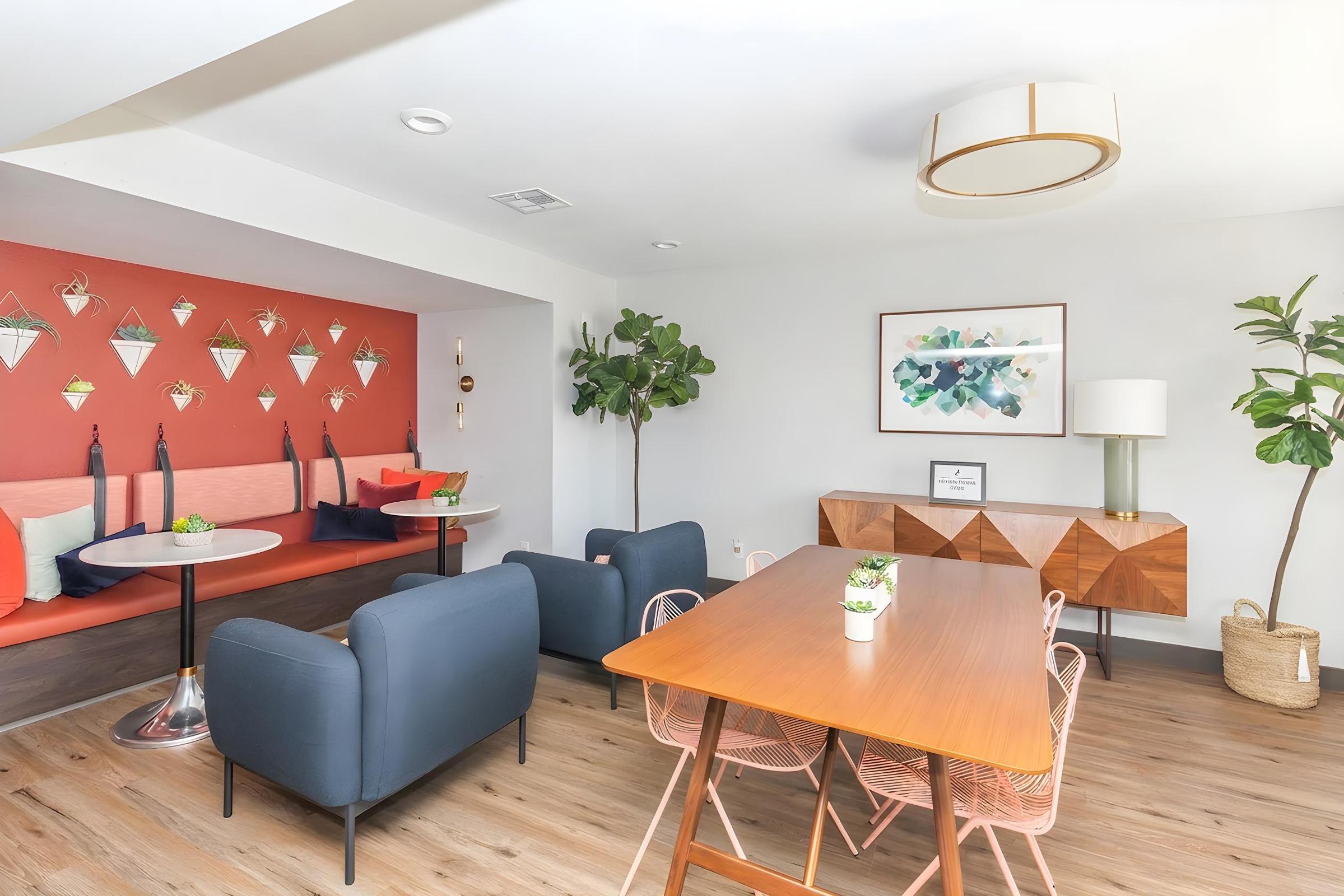 Modern living space featuring a dining area with a wooden table and pink metal chairs, a cozy seating nook with a coral wall and decorative plants, framed artwork on the wall, and stylish light fixtures, creating an inviting atmosphere.