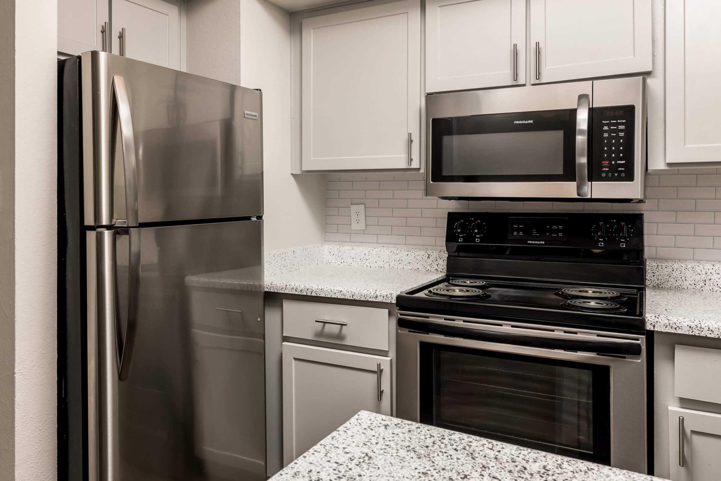 Modern kitchen featuring stainless steel refrigerator and microwave, black stovetop with oven below, and white cabinetry. The countertops are speckled granite, providing a stylish and contemporary look. Bright and clean design with neutral colors.