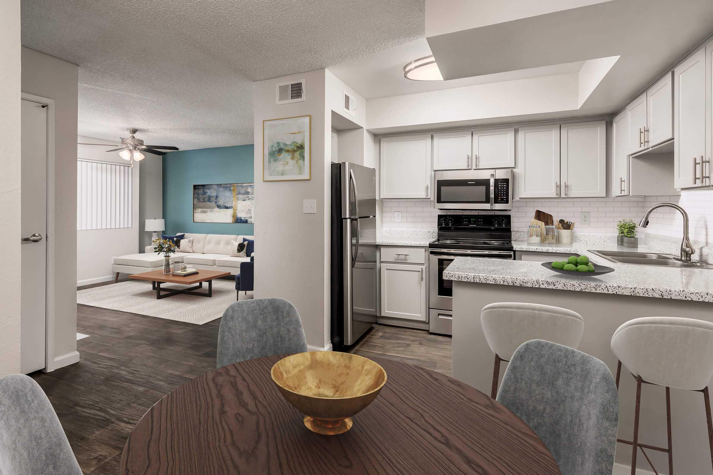 A modern open-concept kitchen and dining area featuring a round wooden table with gray upholstered chairs. The kitchen includes stainless steel appliances, granite countertops, and a bowl of green fruit. In the background, a cozy living room with a light-colored sofa and decorative artwork on a blue wall.