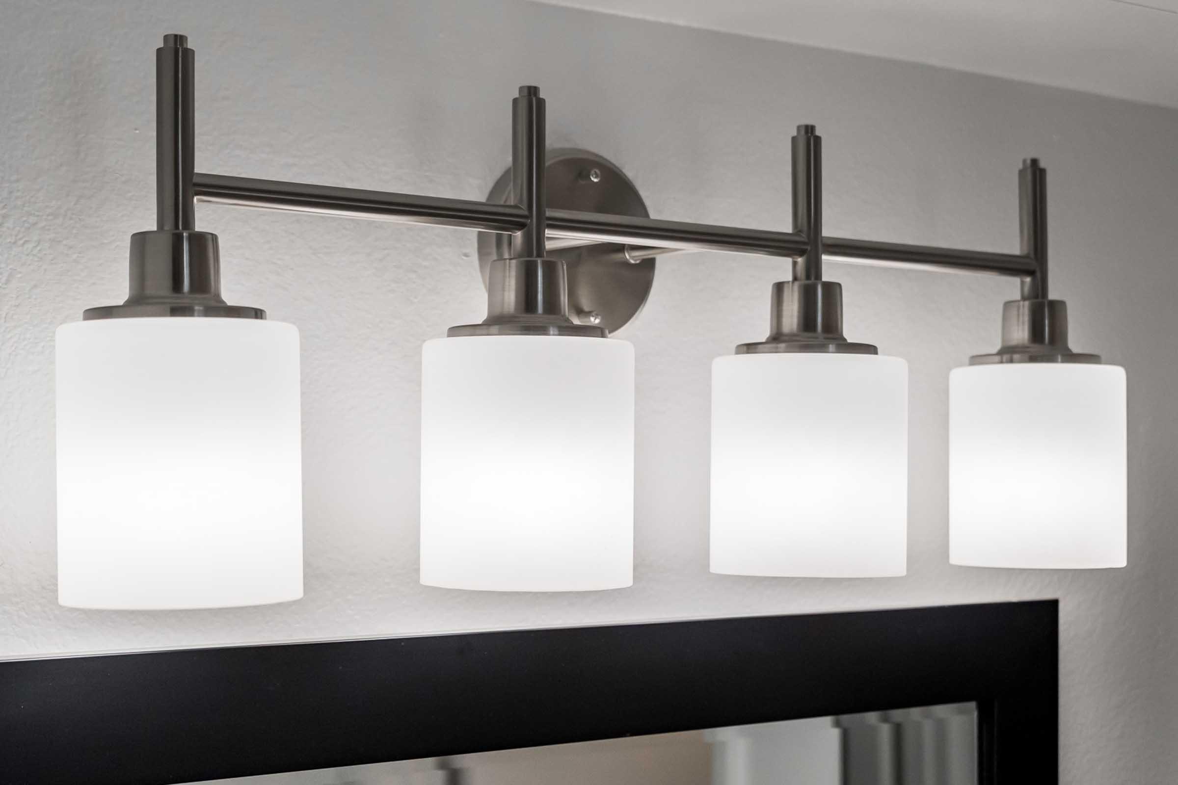 A modern four-bulb wall light fixture with frosted glass shades, mounted on a metal bracket. The fixture is positioned above a mirror, providing soft, even illumination in a bathroom or vanity setting. The design features a sleek and contemporary aesthetic.