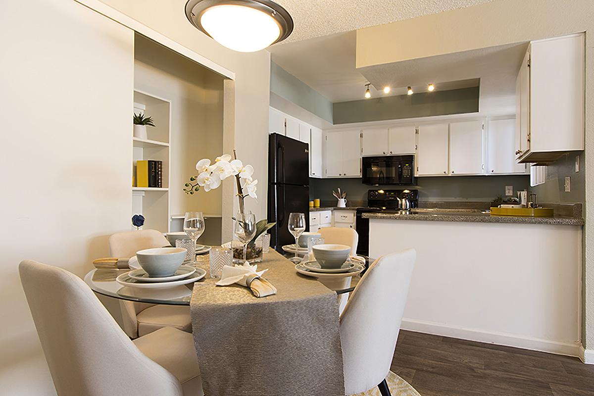 A modern dining area featuring a round glass table set for four with stylish dishware and a table runner. In the background, a well-equipped kitchen with white cabinets and dark appliances. Soft lighting and neutral decor create a warm atmosphere.