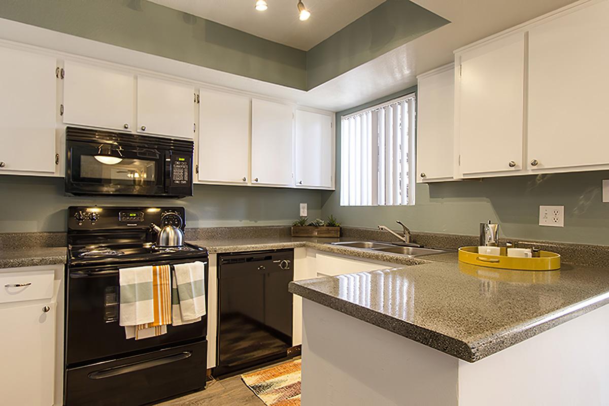 Modern kitchen with white cabinetry, black appliances, and a granite countertop. The layout features a stove, oven, dishwasher, and sink. Natural light comes through a window with blinds. Colorful dish towels hang on the stove, and a small decorative tray sits near the sink.