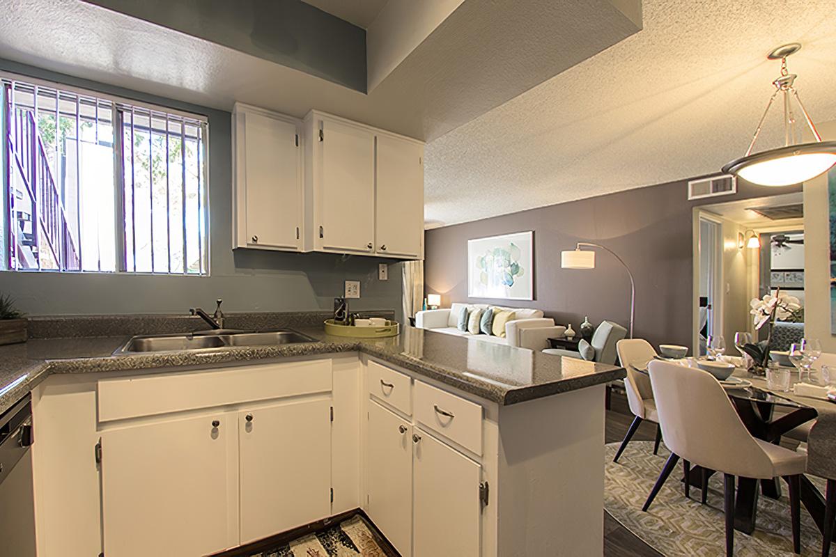 A modern kitchen view featuring white cabinetry, a double sink, and a countertop. In the background, a cozy living area with a sofa, dining table, and decorative artwork. Natural light streams in through a window, illuminating the space with a warm and inviting atmosphere.