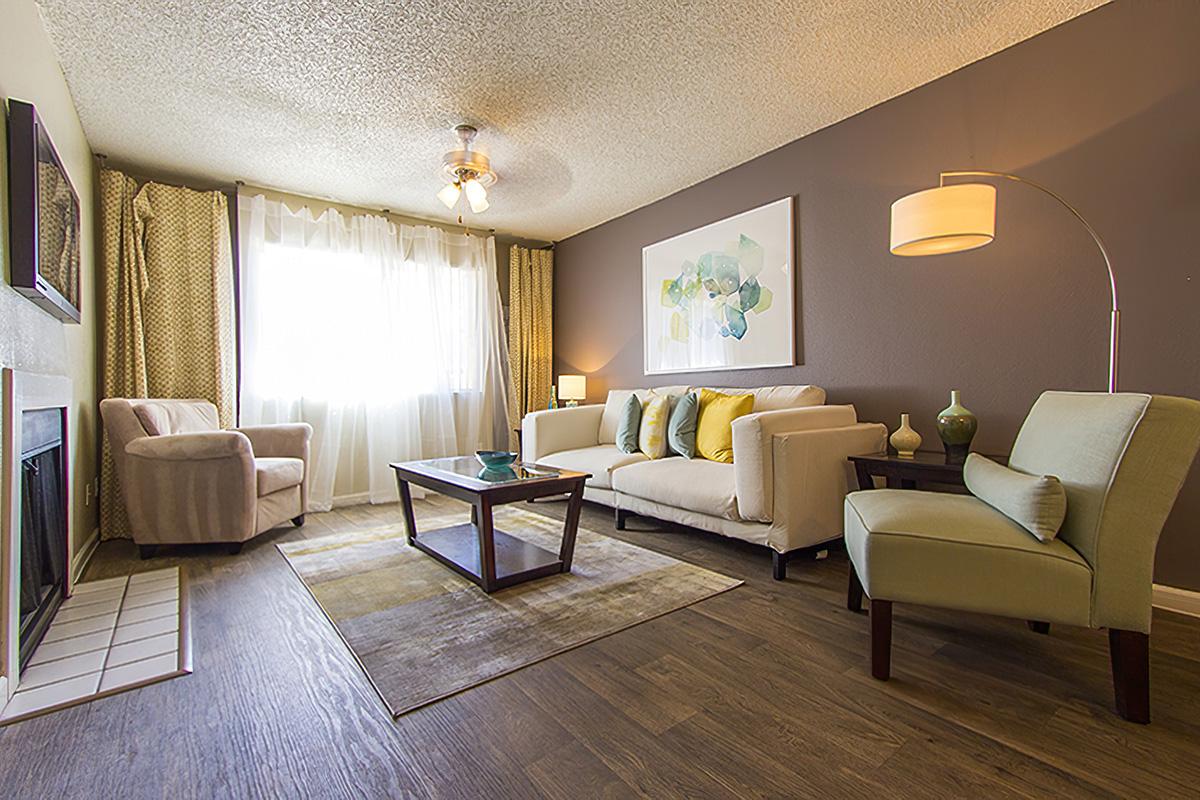 A modern living room featuring a beige sofa with colorful decorative pillows, a glass coffee table, two comfortable chairs, and a light-colored rug. Natural light filters through sheer curtains, with a ceiling fan overhead and a contemporary lamp adding ambiance. A painting hangs on the wall, enhancing the decor.