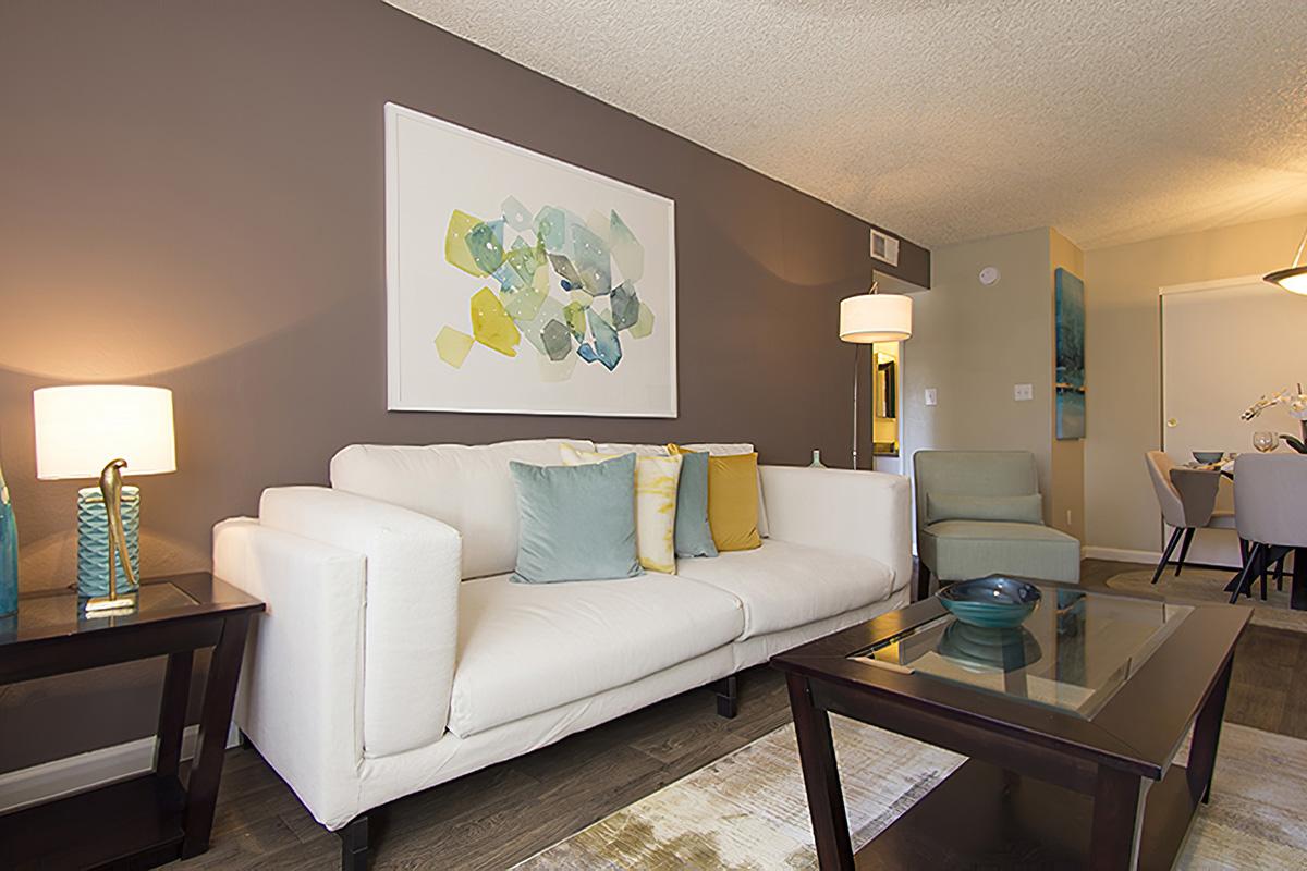 A modern living room featuring a white sofa with colorful pillows, two lamps, a coffee table, and an accent chair. A large abstract artwork in shades of blue and green hangs on the wall, complemented by a neutral brown-painted wall and wooden flooring.