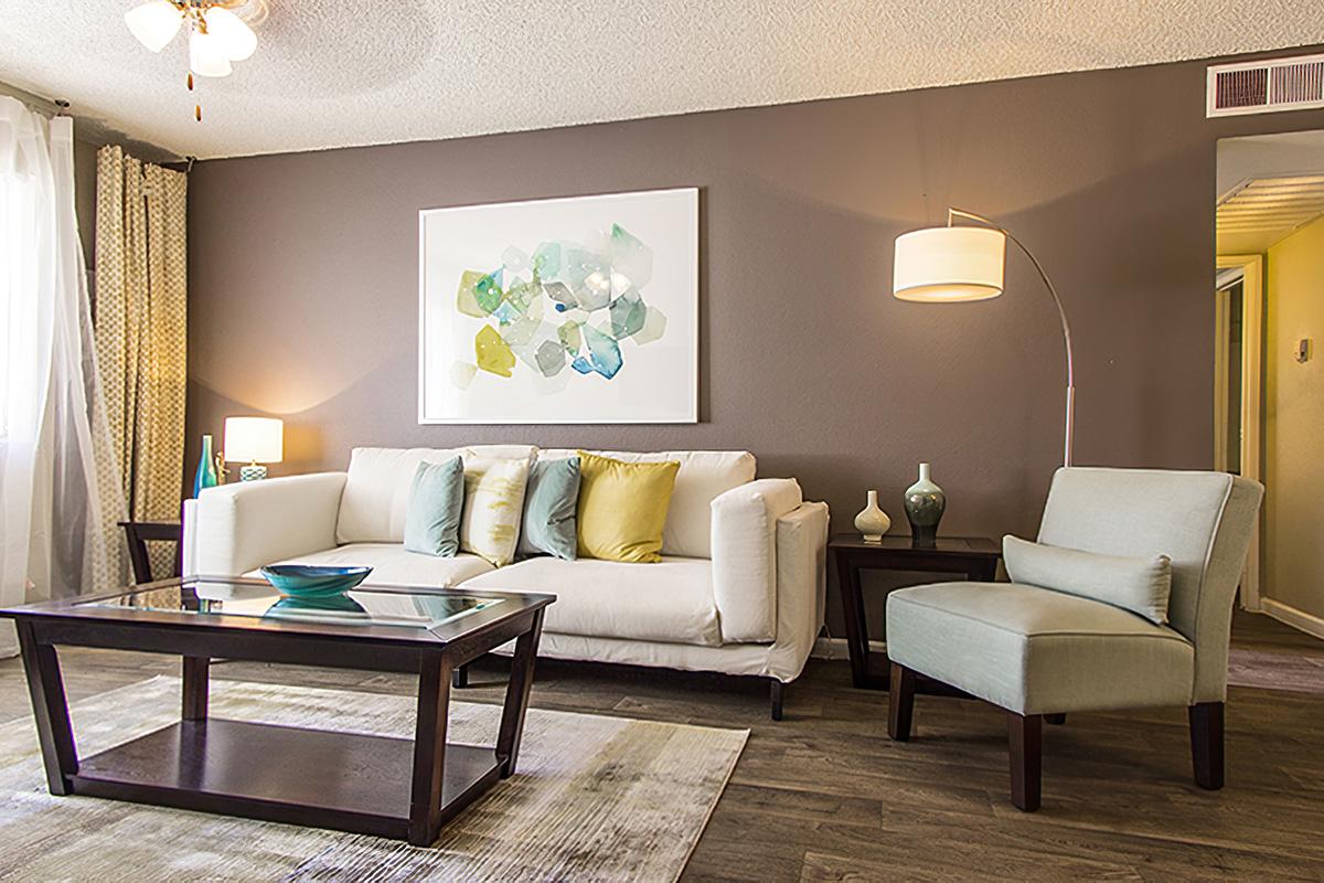 A modern living room featuring a light-colored sofa with colorful pillows, a glass-top coffee table, and a soft armchair. The walls are painted in a warm brown tone, adorned with a large abstract art piece. Soft lighting comes from a floor lamp and there are decorative items arranged on the table and shelf.