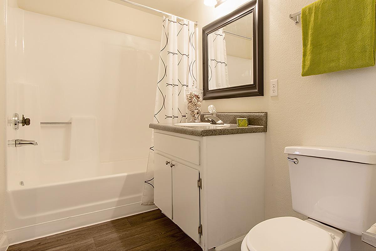 A clean and bright bathroom featuring a bathtub with a shower curtain, a white vanity with a countertop, a mirror above, a green towel hanging on the wall, and a white toilet. The flooring is dark, creating a contrast with the light-colored walls.