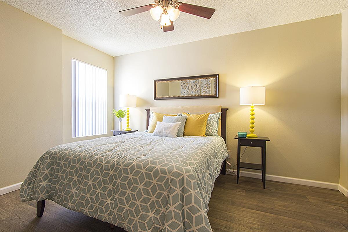 A cozy bedroom featuring a bed with a light blue and white geometric patterned quilt, yellow and white accent pillows, two bedside tables with bright yellow and white lamps, and a decorative mirror above the headboard. Natural light streams in through a window. The walls are painted a soft beige.