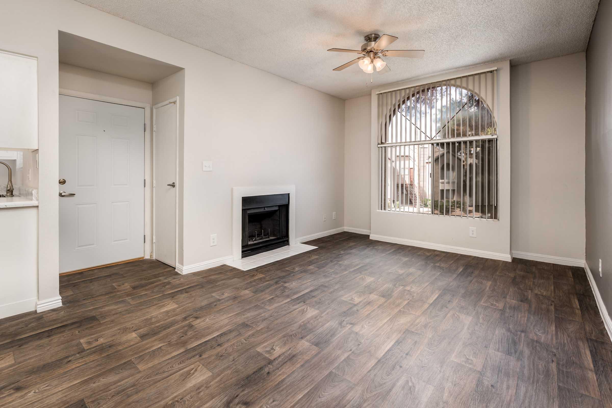A spacious living room with wooden laminate flooring, a ceiling fan, and large windows with blinds. A decorative fireplace is located on one wall, and there is a doorway leading to an entrance. The walls are painted a light color, creating a bright and inviting atmosphere.