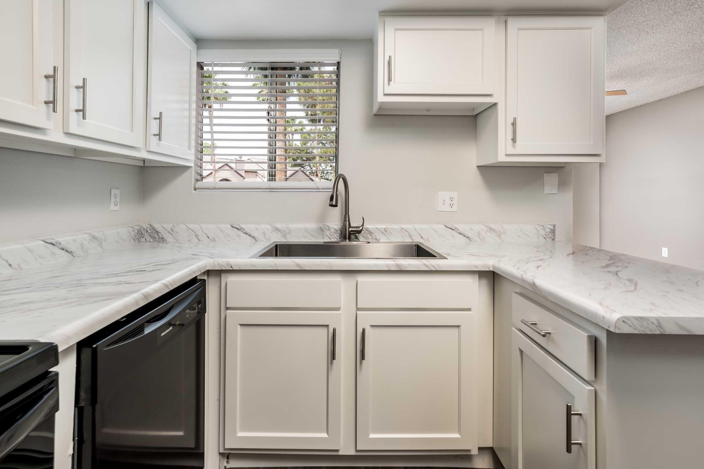 Modern kitchen with white cabinets and a marble countertop. The sink is positioned in the corner under a window with horizontal blinds. A black dishwasher is visible beside the sink, and the room features light-colored walls and a ceiling with a light fixture.