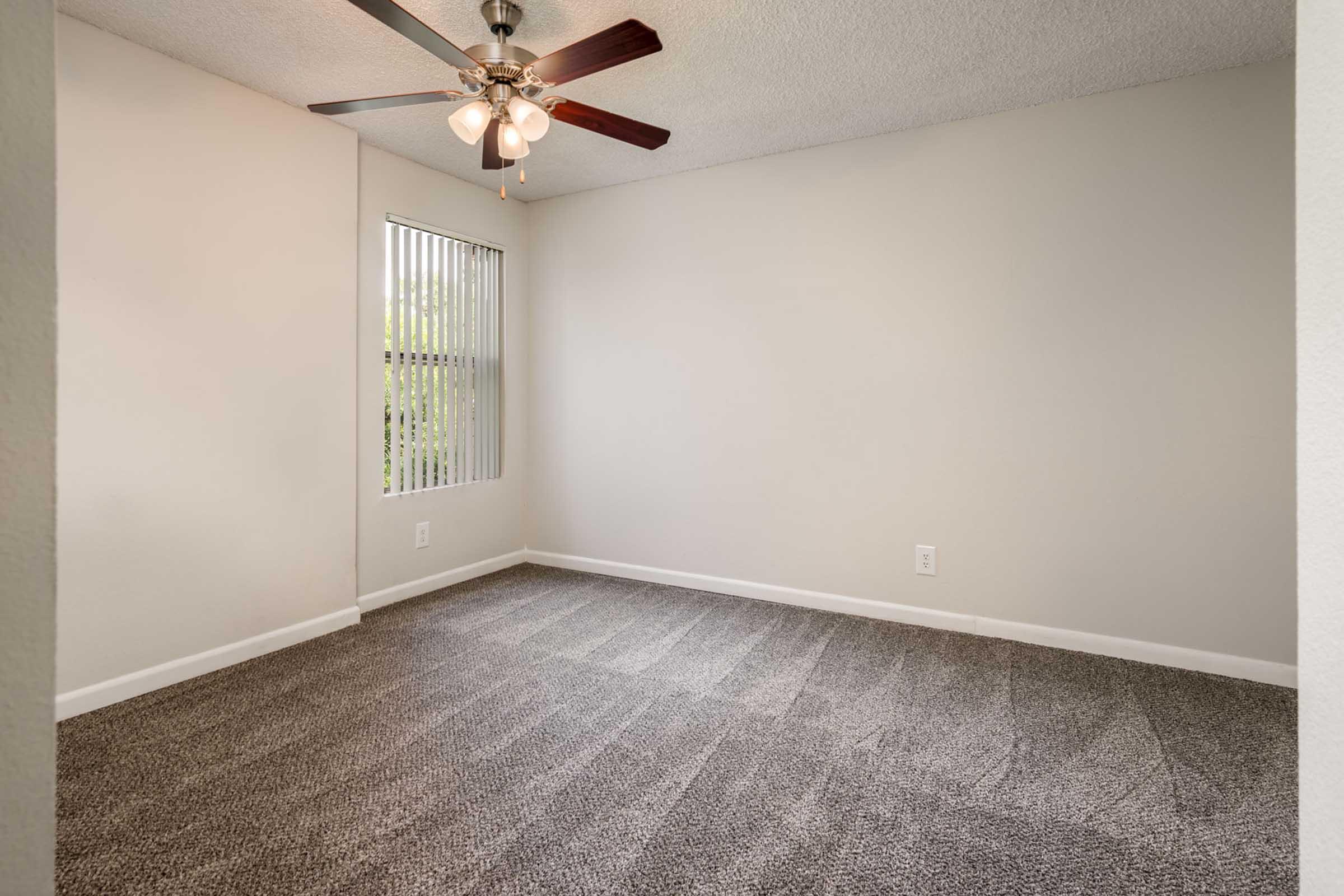 Empty room with light gray carpet and beige walls. A ceiling fan with four bulbs hangs in the center, and there's a window with vertical blinds allowing natural light to enter. The space is well-lit and clean, suitable for various uses such as a bedroom or office.