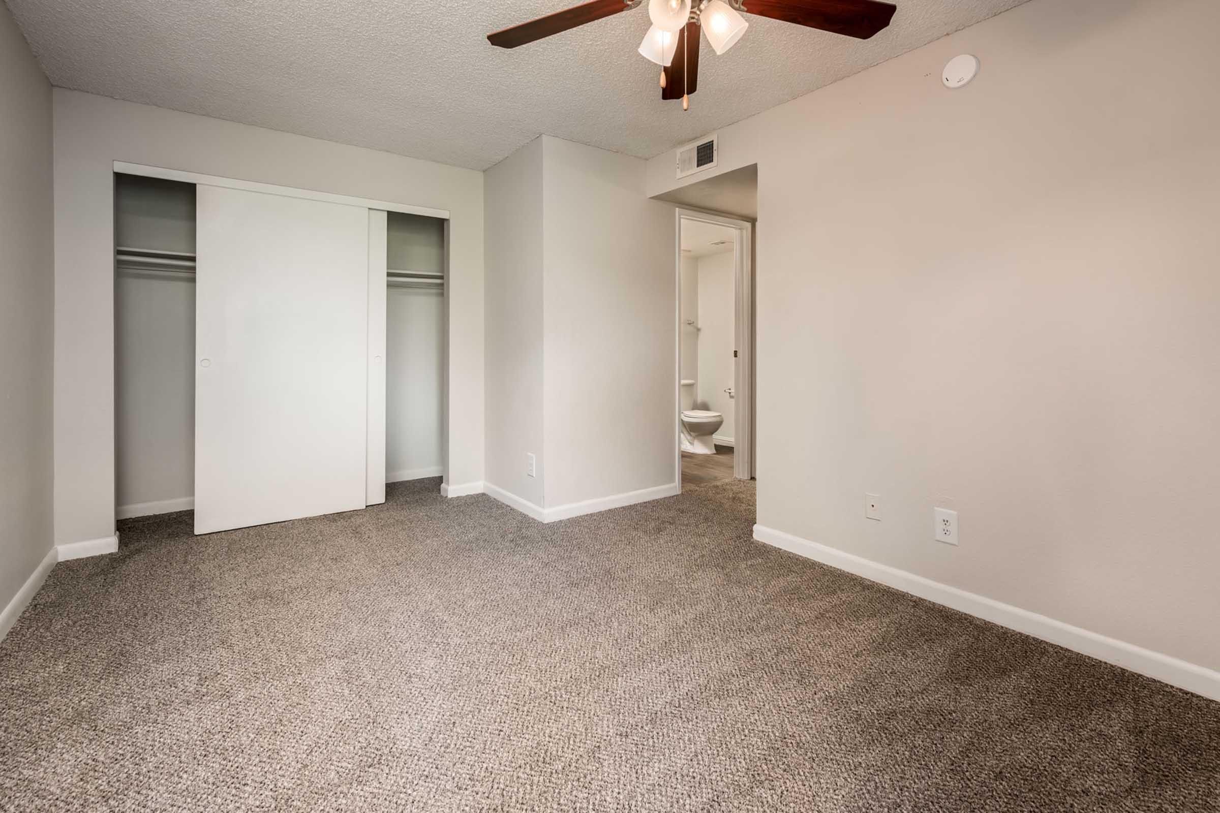 A spacious empty bedroom with beige carpet, a ceiling fan with light, and a closet with sliding doors. A door leads to a bathroom in the background, enhancing the room's layout. The walls are painted in a neutral tone, offering a bright and airy atmosphere.