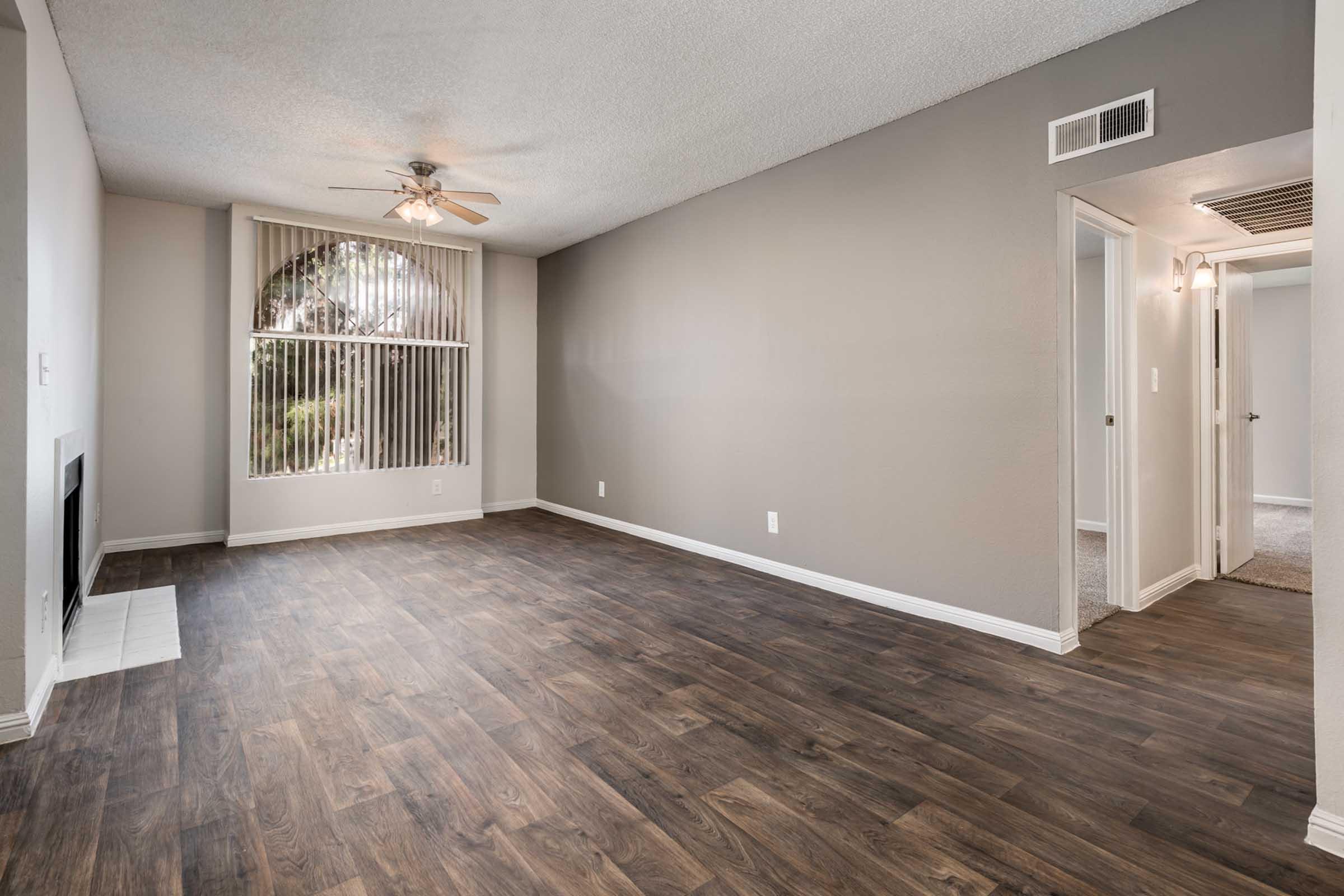 A spacious living room with a ceiling fan and large window featuring vertical blinds. The floor is made of dark wood, and there is a white fireplace to the left. The walls are painted in a light gray color, and there are doorways leading to other rooms. Natural light fills the space, giving it a bright appearance.
