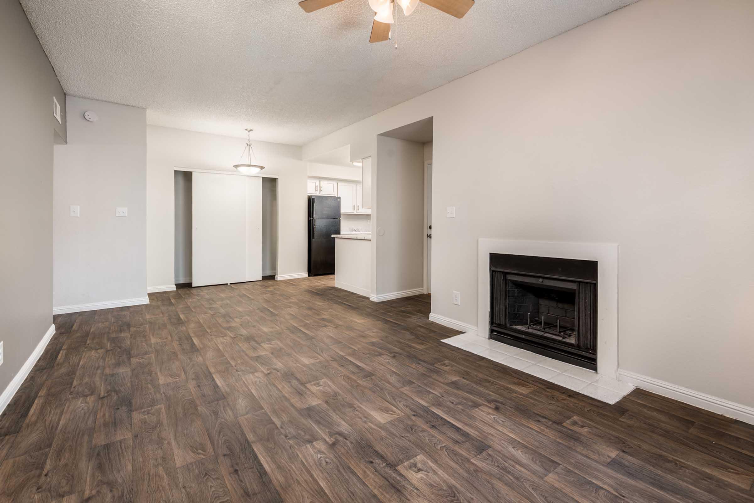 A spacious living area with wooden flooring, featuring a ceiling fan and a fireplace. In the background, there's a kitchen area with light-colored cabinetry, a door leading to a hallway, and a door to a closet. Natural light enhances the warm, neutral tones of the walls.