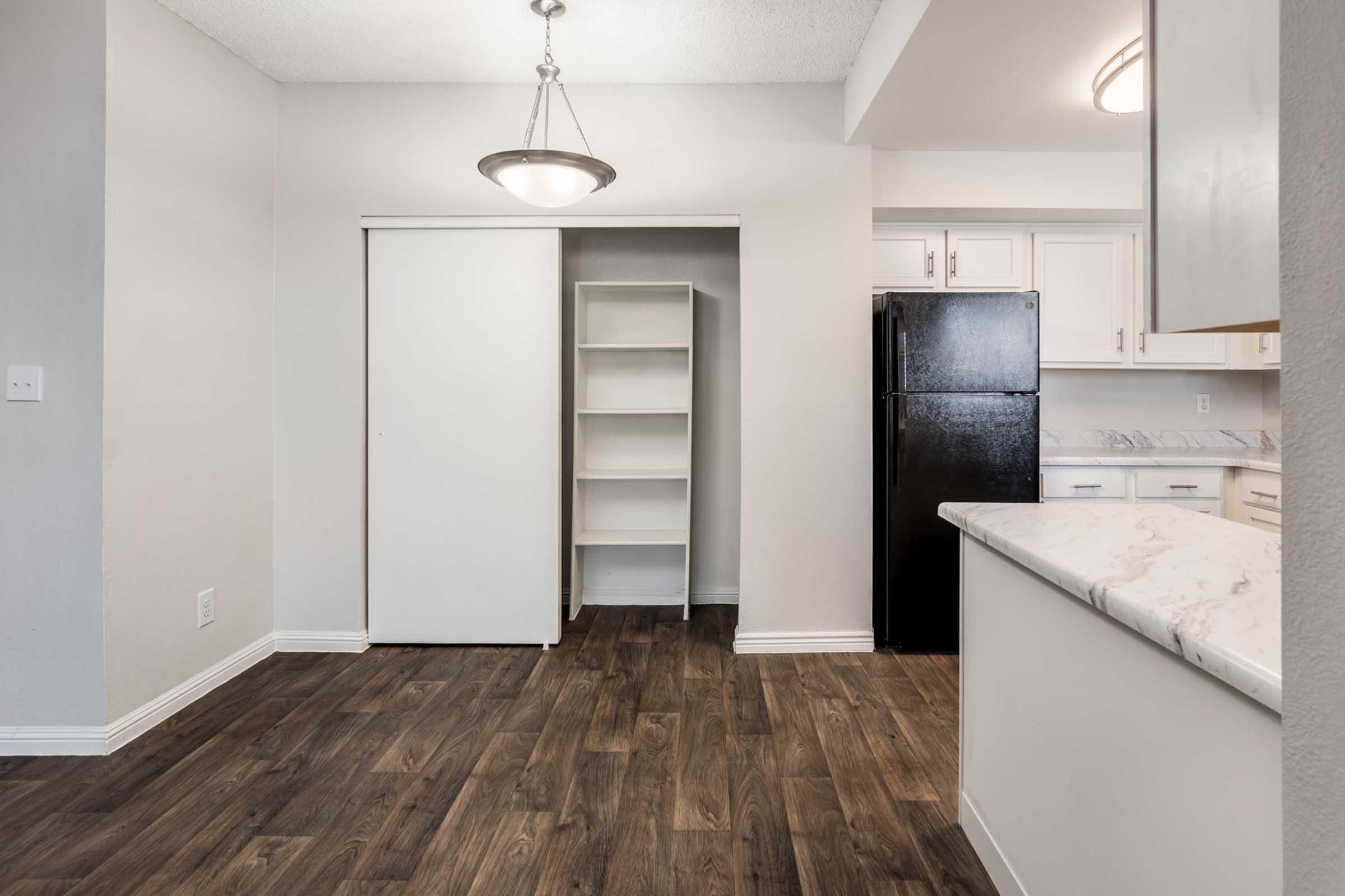 A modern kitchen featuring a black refrigerator, white cabinetry, and a light fixture. There's a pantry or storage closet on the left and a countertop view with a marble finish. The floor is a warm-toned wood laminate, creating a cozy atmosphere in the space.