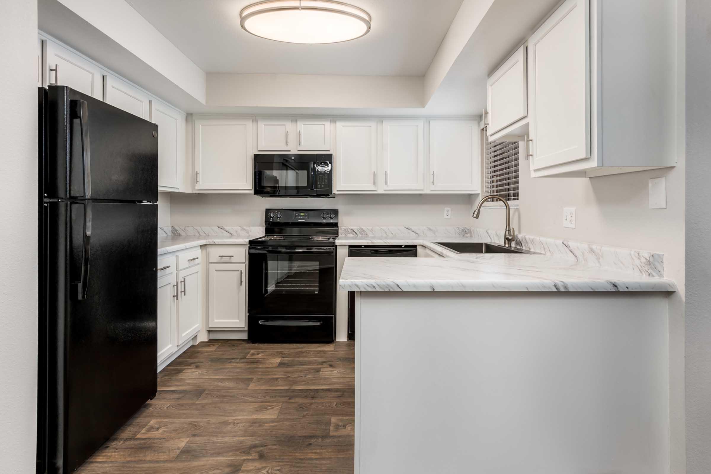 Modern kitchen featuring white cabinetry, marble countertops, a black refrigerator, and a black microwave and oven. The space is well-lit with a circular ceiling light, and there is a sink integrated into the countertop. The flooring is wood-like, adding warmth to the contemporary design.