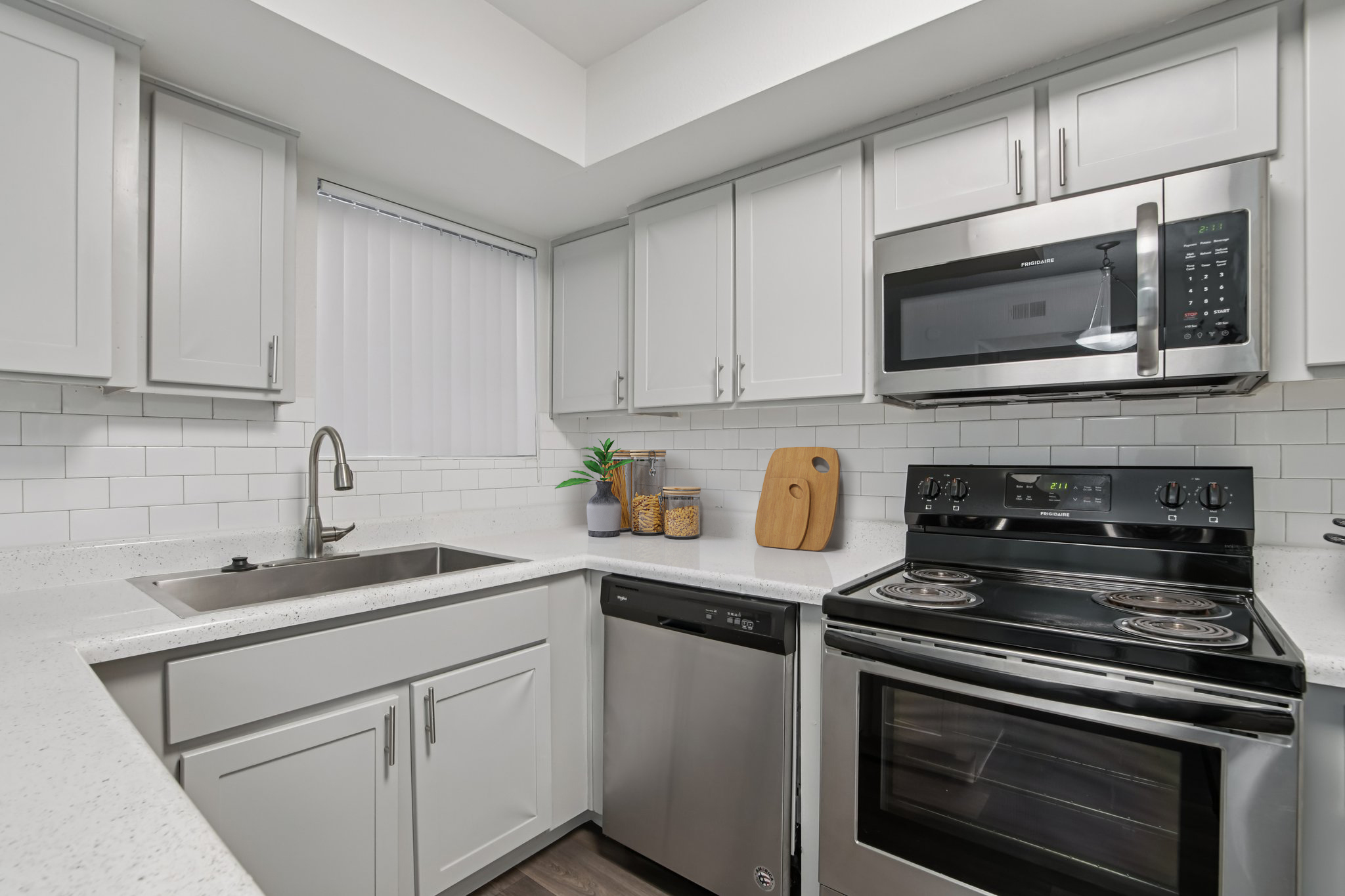 Modern kitchen with white cabinetry, a stainless steel sink, and appliances including a dishwasher and microwave. Countertops are light-colored with a textured finish. A small wood cutting board and a plant add a touch of decor. The space has a bright and clean aesthetic.