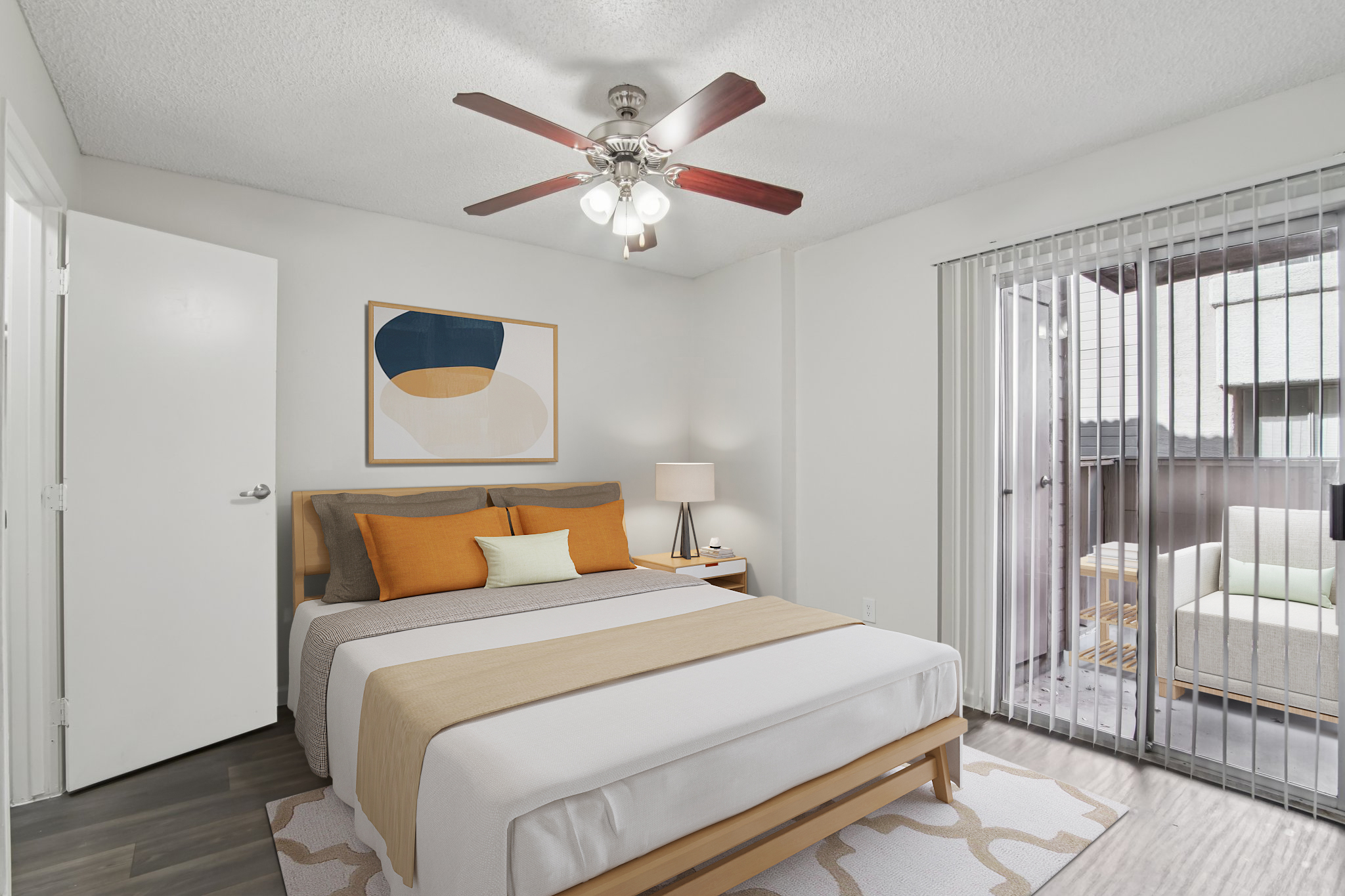 A modern bedroom featuring a neatly made bed with orange and beige accents, a ceiling fan, and a bedside lamp. A piece of abstract art hangs on the wall above the bed. Large sliding glass doors lead to a patio area with outdoor seating. The room has light-colored walls and wooden flooring.