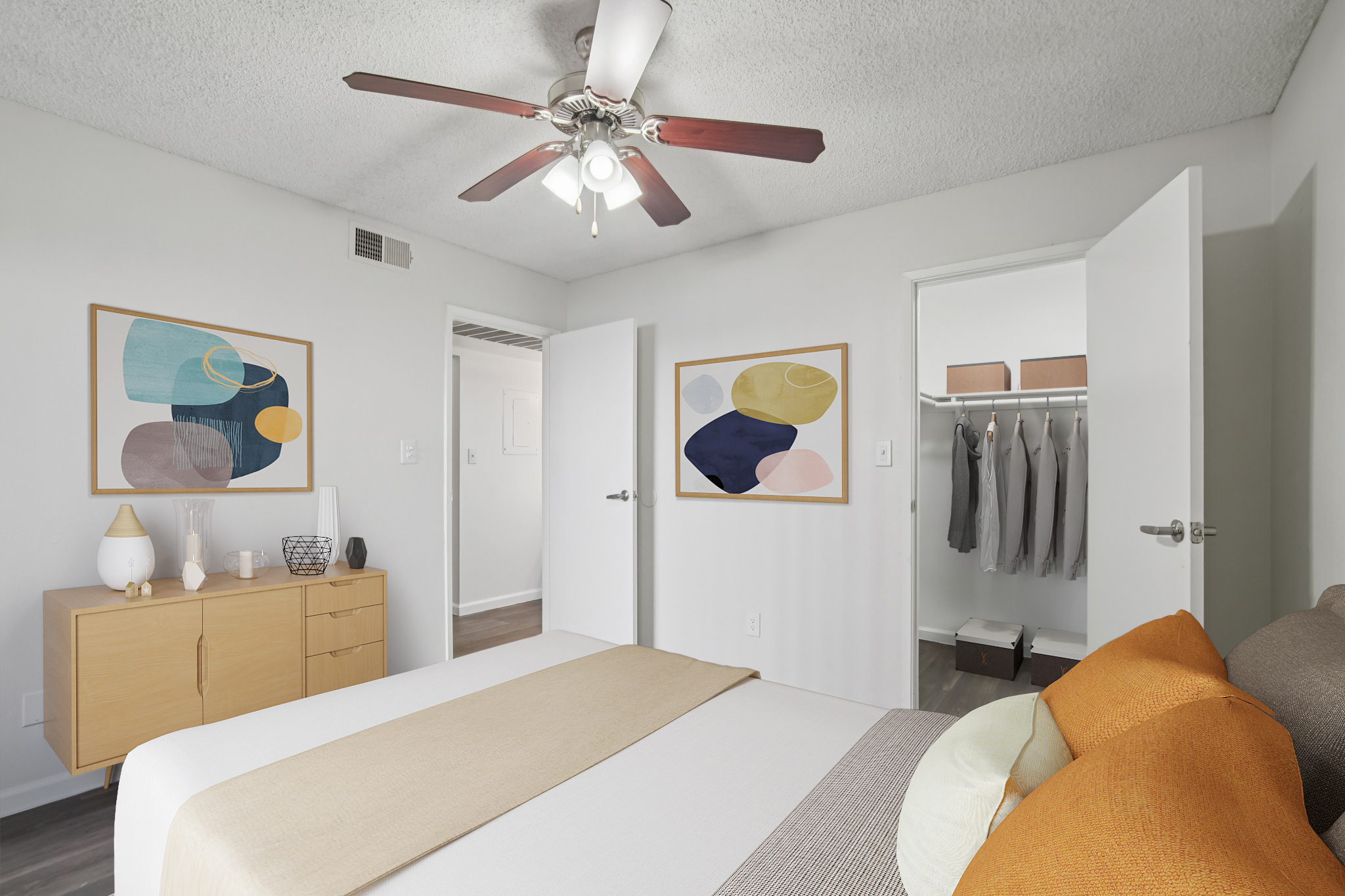 A well-decorated bedroom featuring a bed with colorful pillows, a wooden nightstand, and abstract art on the walls. A ceiling fan hangs above, and a closet with neatly arranged clothes is visible through an open door. The room has a clean, modern aesthetic with light-colored walls and hardwood flooring.