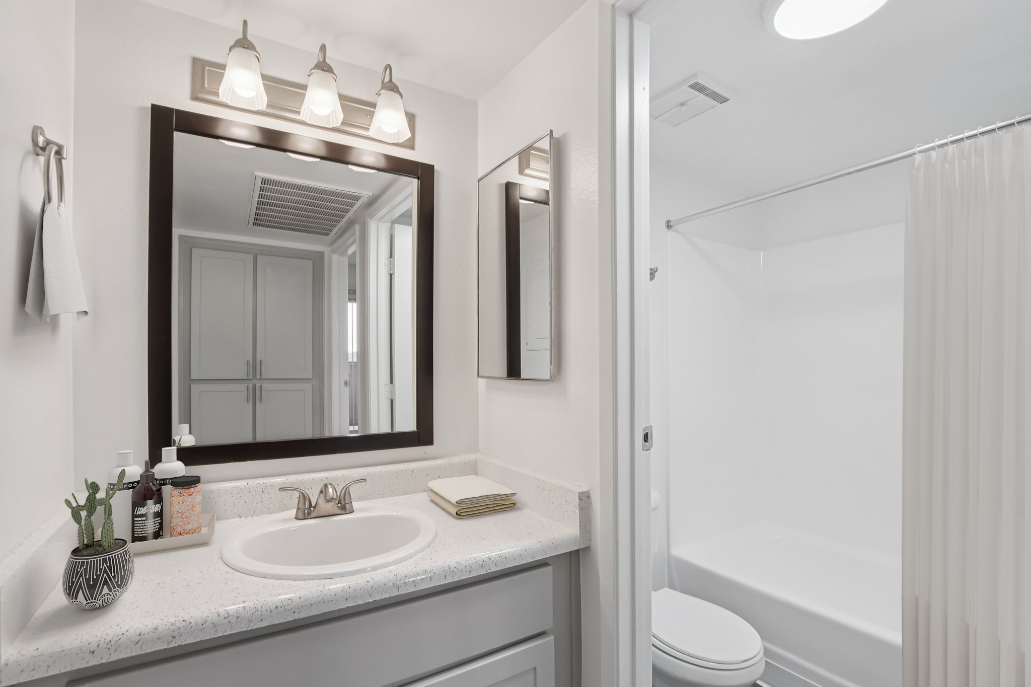Modern bathroom featuring a white vanity with a round sink, a large mirror with decorative lighting, a shower area with a curtain, and neutral-colored walls. The countertop is adorned with toiletries and a small decorative plant, creating a clean and minimalistic aesthetic.