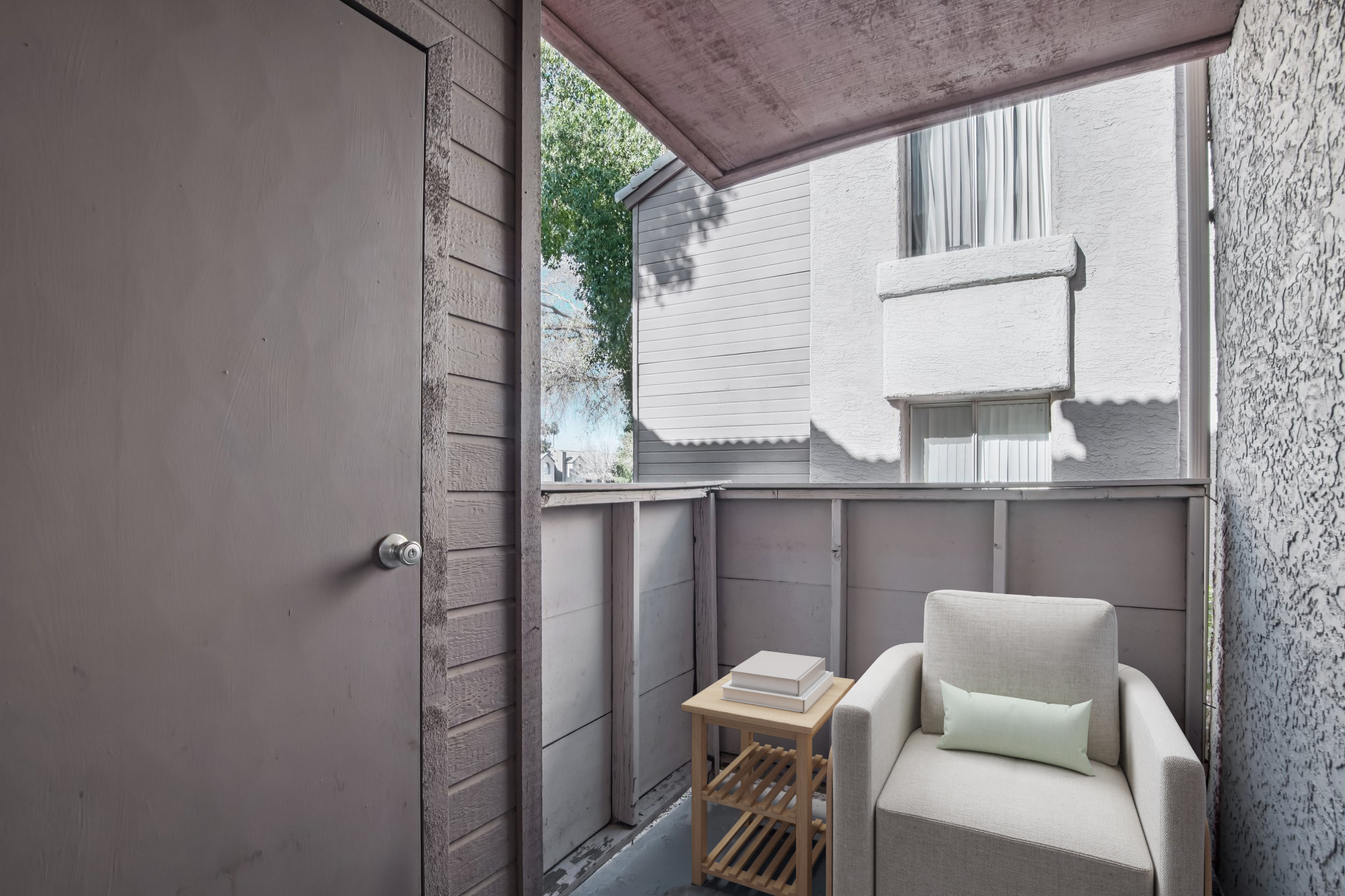 A small balcony view featuring a light-colored chair with a soft green cushion, a wooden side table with a stack of books, and a door leading inside. The scenery includes a glimpse of greenery outside and a neighboring building in the background, creating a cozy outdoor space.