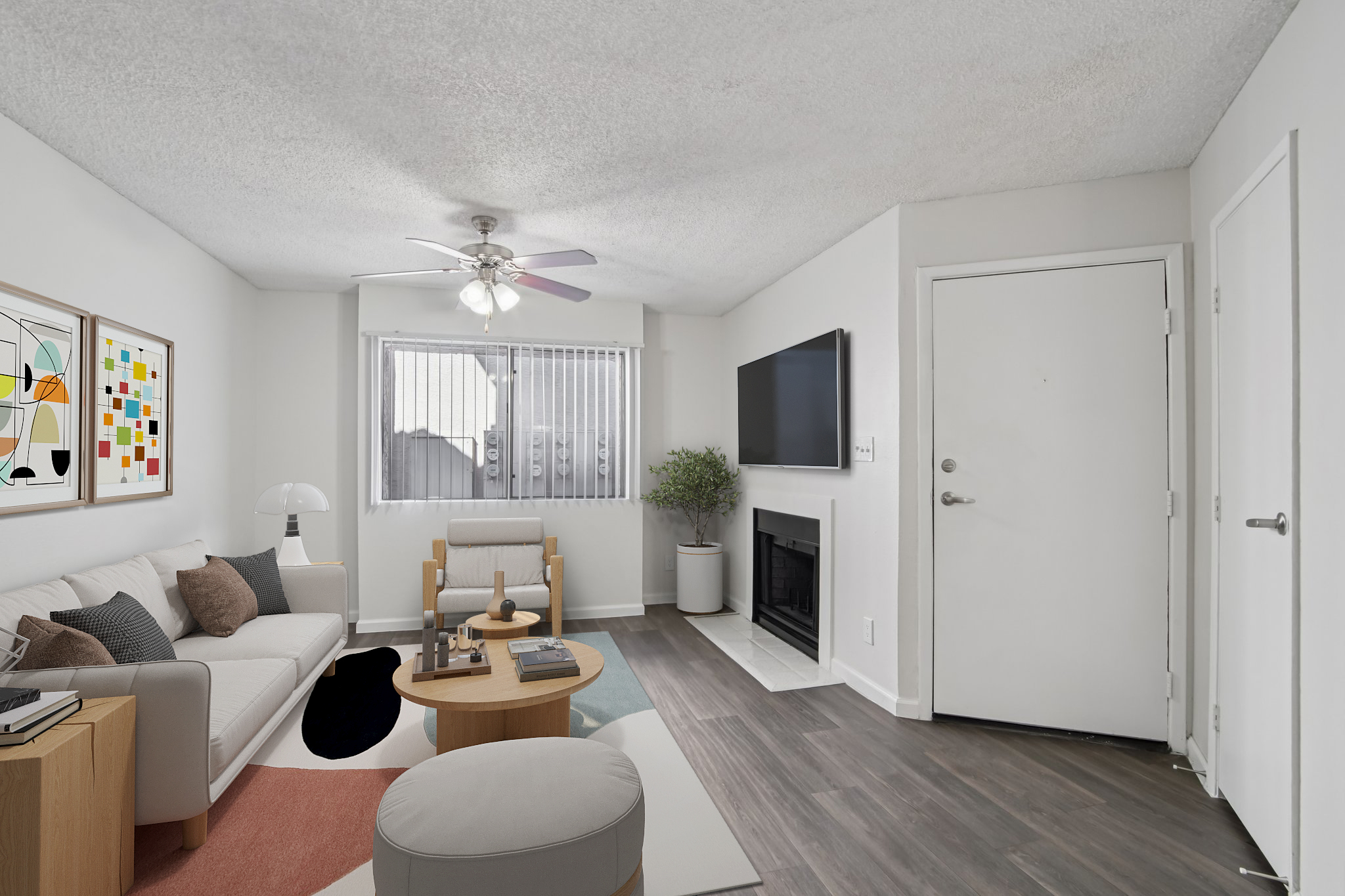 A modern living room featuring a light-colored sofa, a small round coffee table, and a lounge chair. A television is mounted on the wall, and a fireplace is visible. Natural light enters through a window with blinds. The decor includes artwork on the walls and a potted plant for a cozy ambiance.