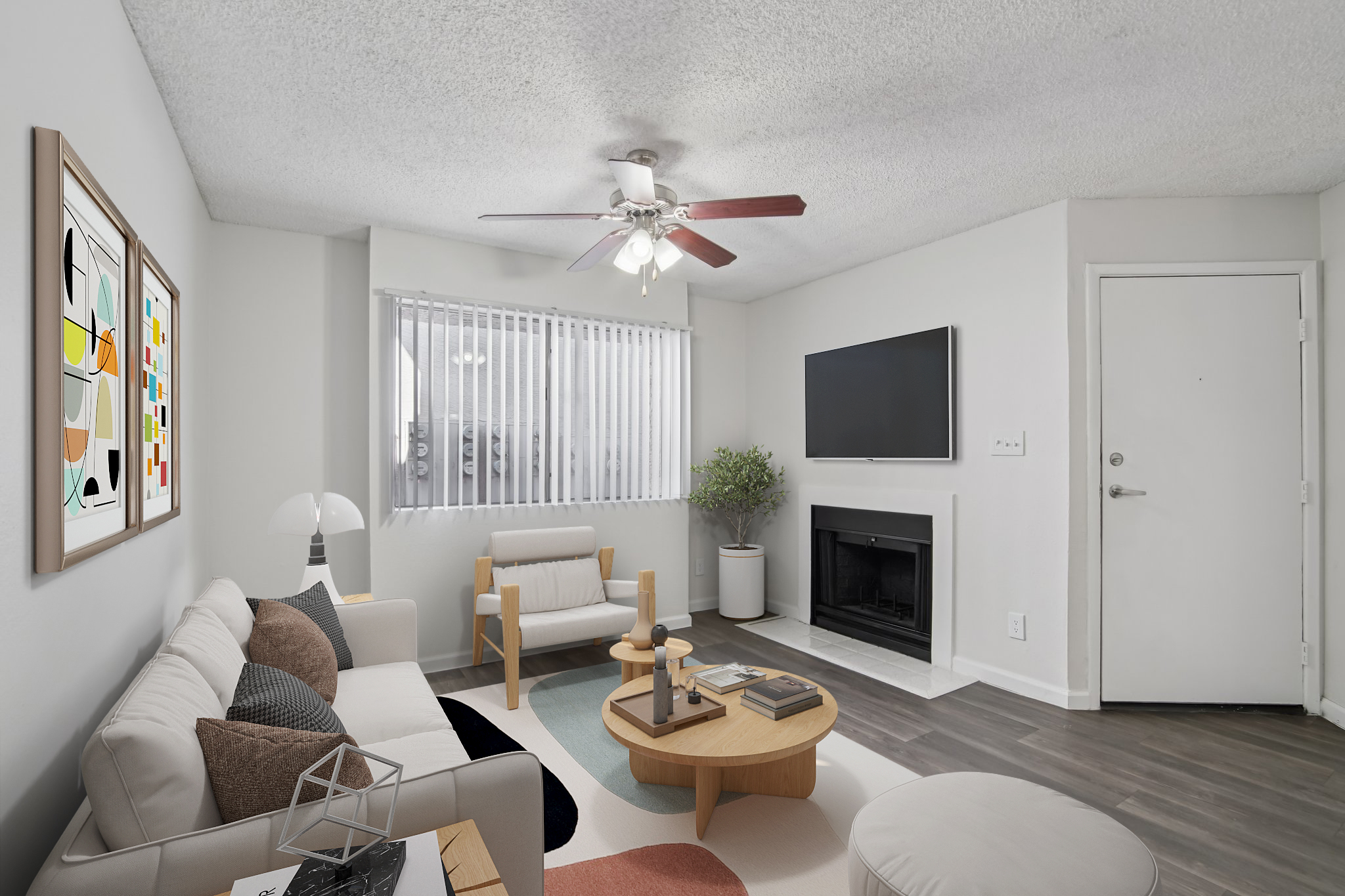 A modern living room featuring a light-colored sofa, a wood coffee table, and a TV mounted on the wall. There is a ceiling fan with three lights, a decorative area rug, and a small indoor plant. Window blinds allow natural light, and artwork is displayed on one wall, adding a vibrant touch to the space.