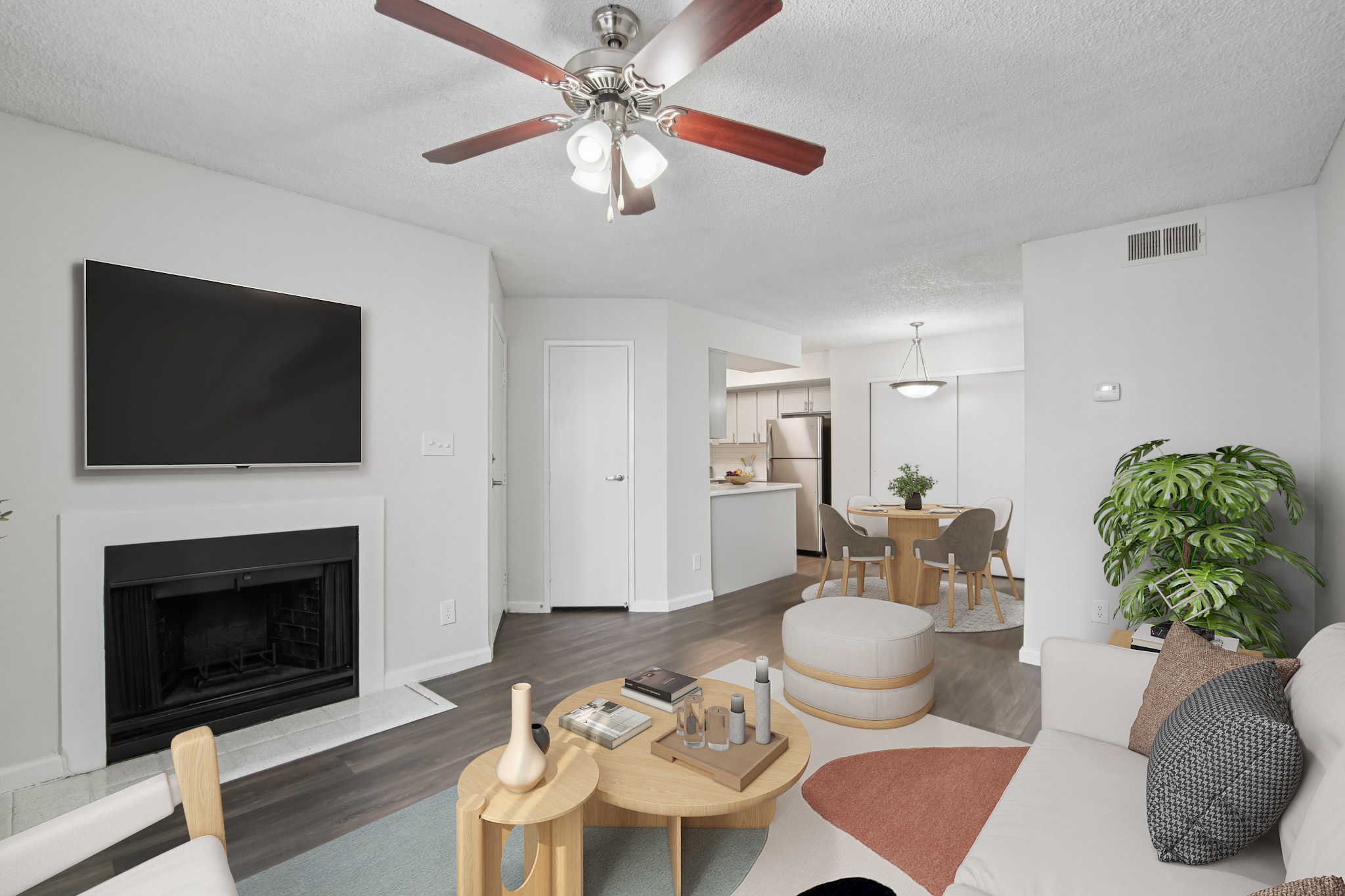 A modern living room featuring a light-colored sofa, a wooden coffee table with decorative items, and a ceiling fan. A wall-mounted television and a fireplace are visible, along with a dining area in the background. Natural light brightens the space, complemented by green potted plants.