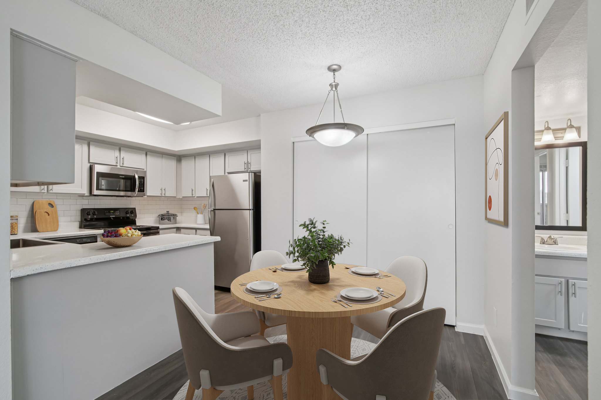 A modern, bright kitchen and dining area featuring a round wooden table with four chairs, a bowl of fruit on the table, stainless steel appliances, and light-colored cabinets. Natural light floods the space, enhancing the clean, contemporary aesthetic.