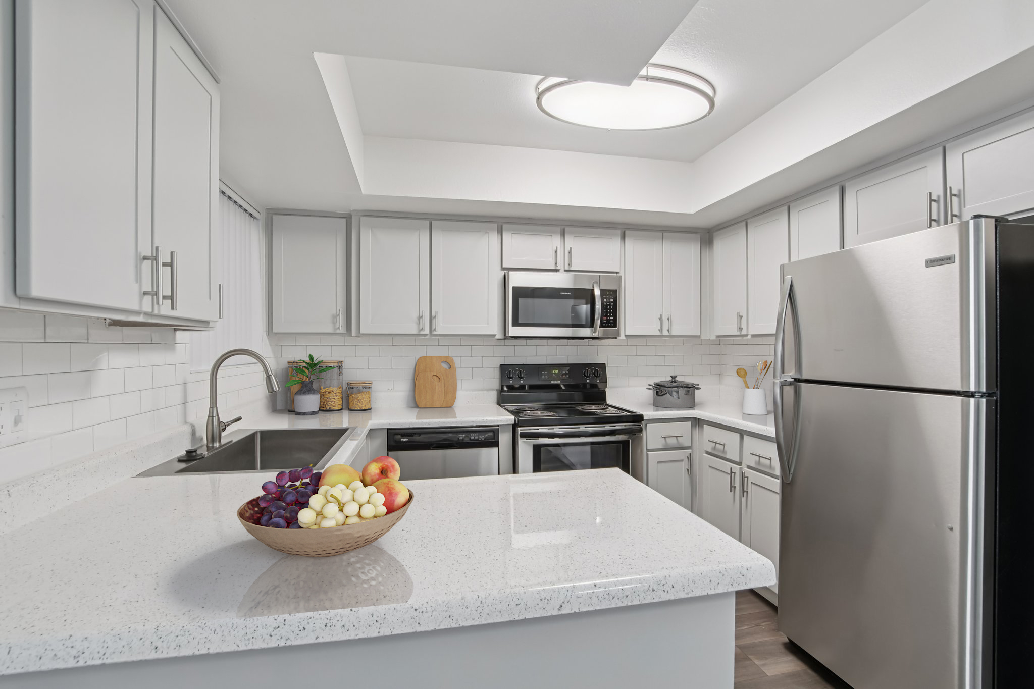 A modern kitchen featuring white cabinetry and a gray countertop. Stainless steel appliances include a microwave, stove, and refrigerator. A wooden cutting board is on the counter beside a bowl of assorted fruits, including grapes, bananas, and apples. The space is well-lit with recessed lighting.