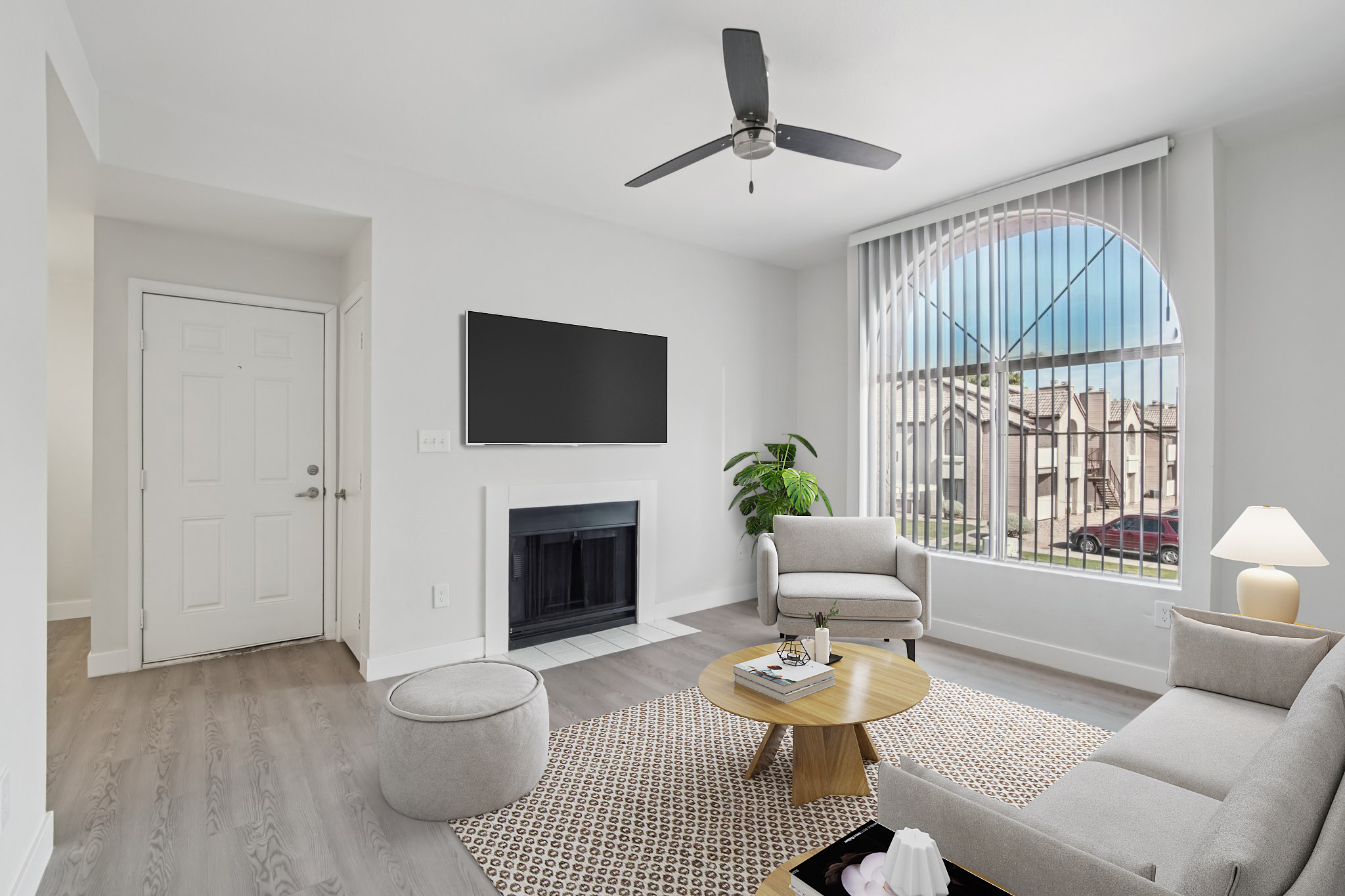A bright and modern living room featuring a light gray color scheme. It includes two beige armchairs, a round ottoman, a coffee table, and a wall-mounted television. There's a fireplace below the TV, a large window with vertical blinds, and a potted plant near the window, creating an inviting atmosphere.