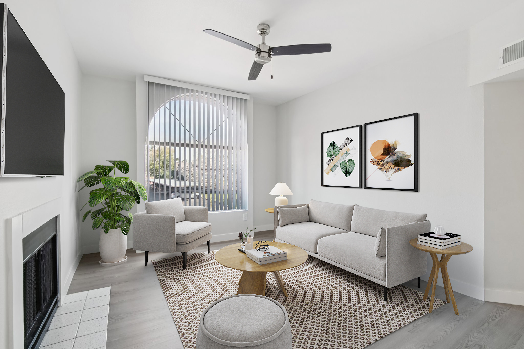 A modern living room featuring a light-colored sofa and armchairs, a round wooden coffee table, and a rug. There are decorative plants, a wall-mounted TV, and two framed artworks on the walls. Bright natural light streams through a large window with vertical blinds, creating a welcoming atmosphere.