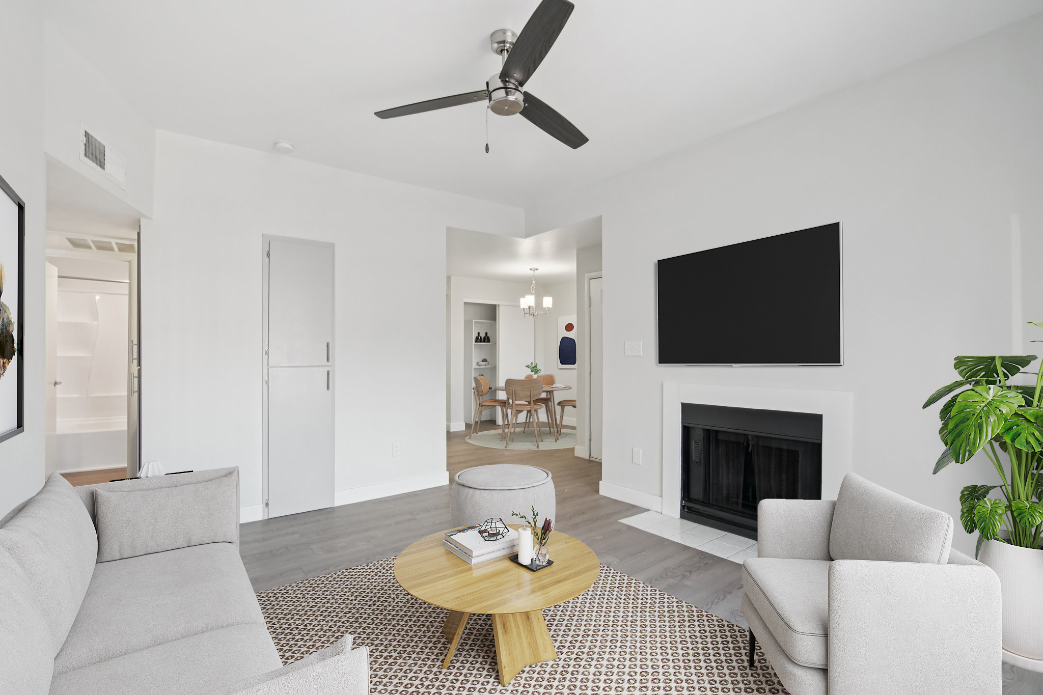 A modern living room featuring a light gray sofa and armchairs around a round wooden coffee table. There is a ceiling fan and a mounted flat-screen TV on the wall. A fireplace is visible, and a large plant adds greenery. The space has neutral colors and a cozy, inviting atmosphere.
